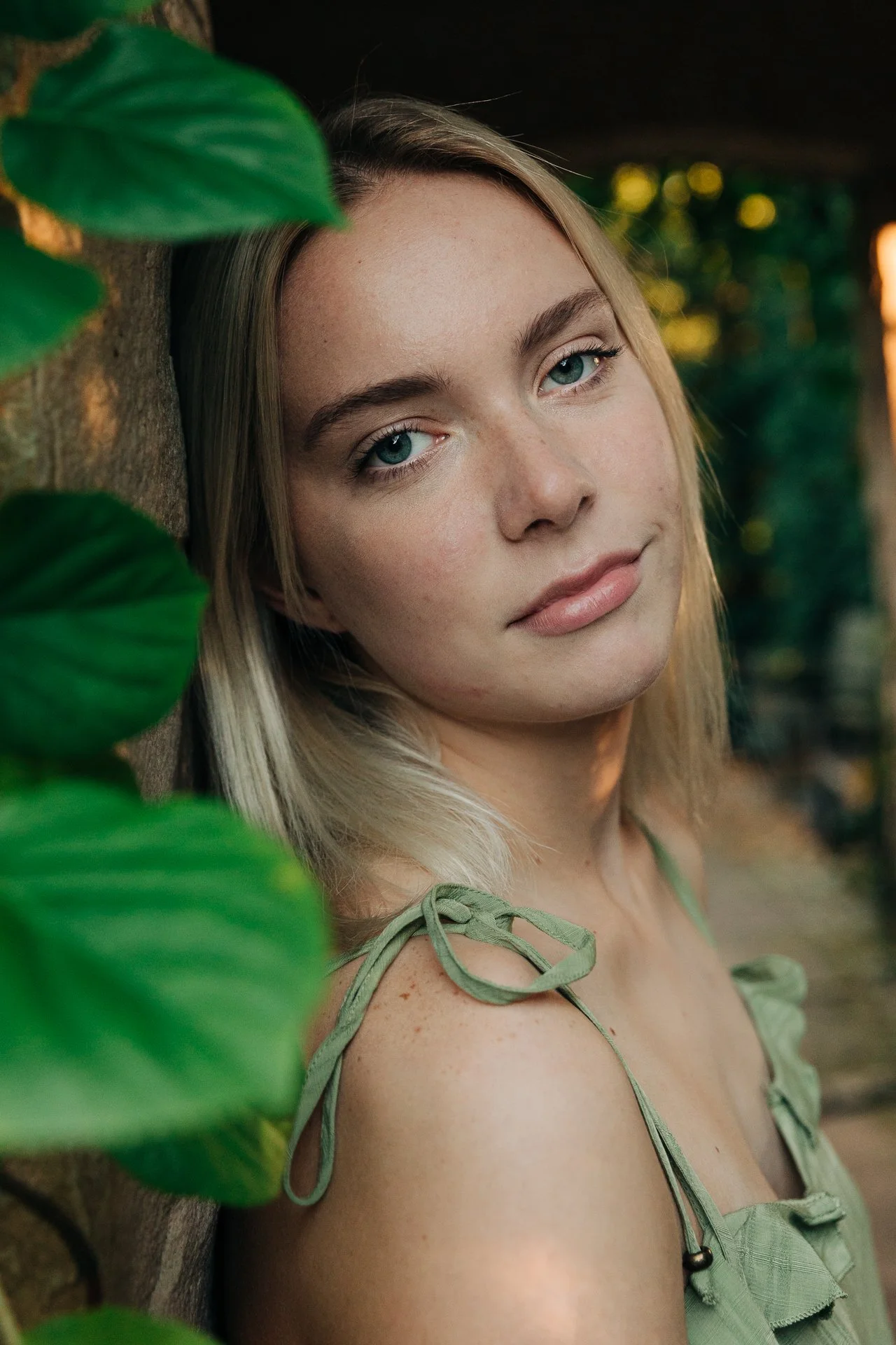 A young woman with blonde hair and blue eyes leaning against a wooden surface, surrounded by green leaves, with a soft natural light in the background.