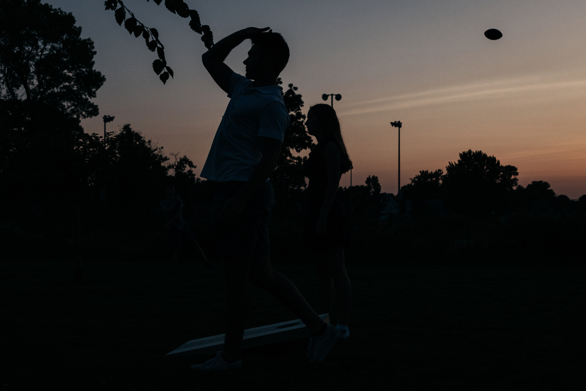 Silhouettes of two people playing cornhole outdoors during sunset, with trees and light poles in the background.