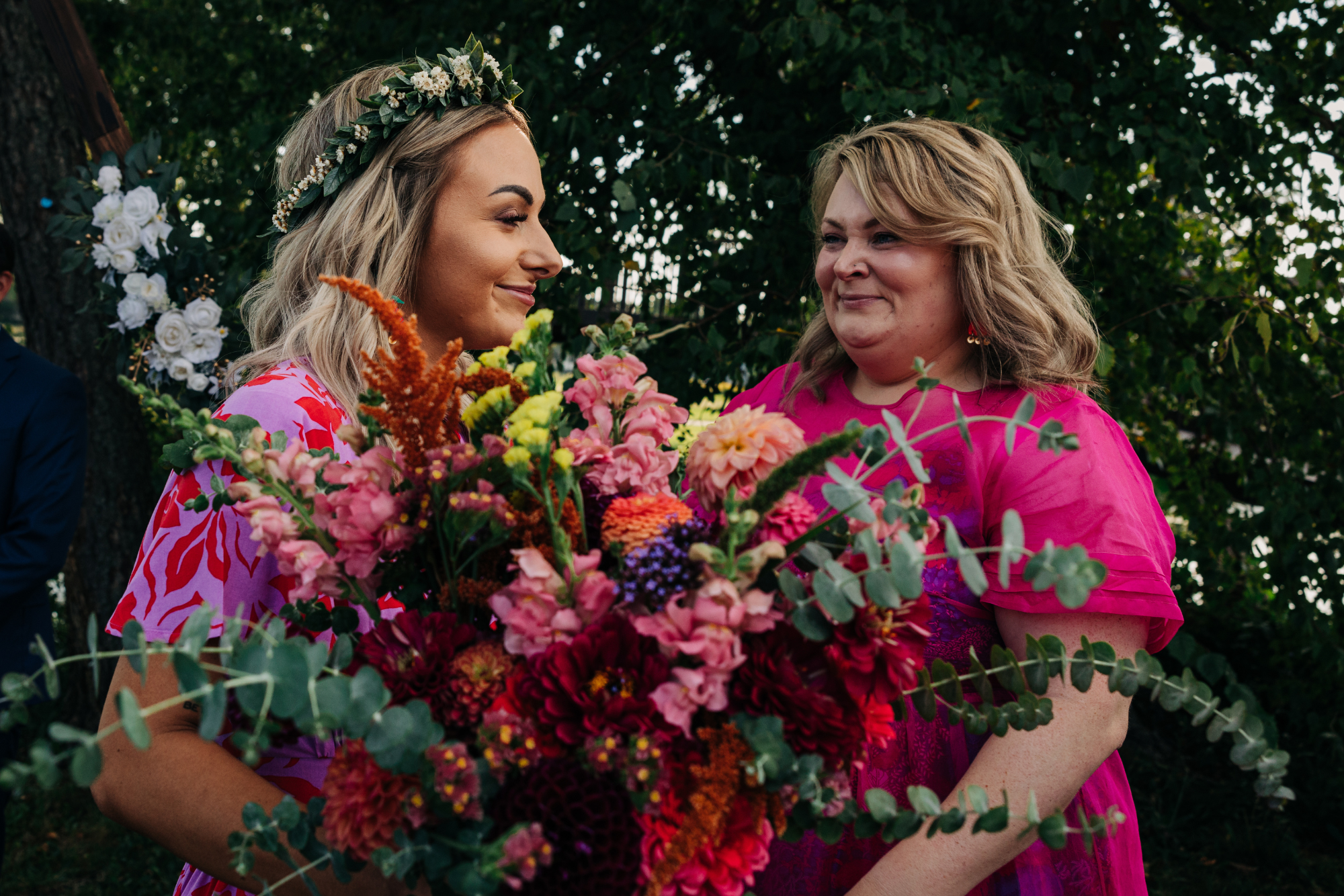 Two women, one with a flower crown, exchanging a kiss at an outdoor wedding, holding a large bouquet of colorful flowers, with a tree and floral decorations in the background.