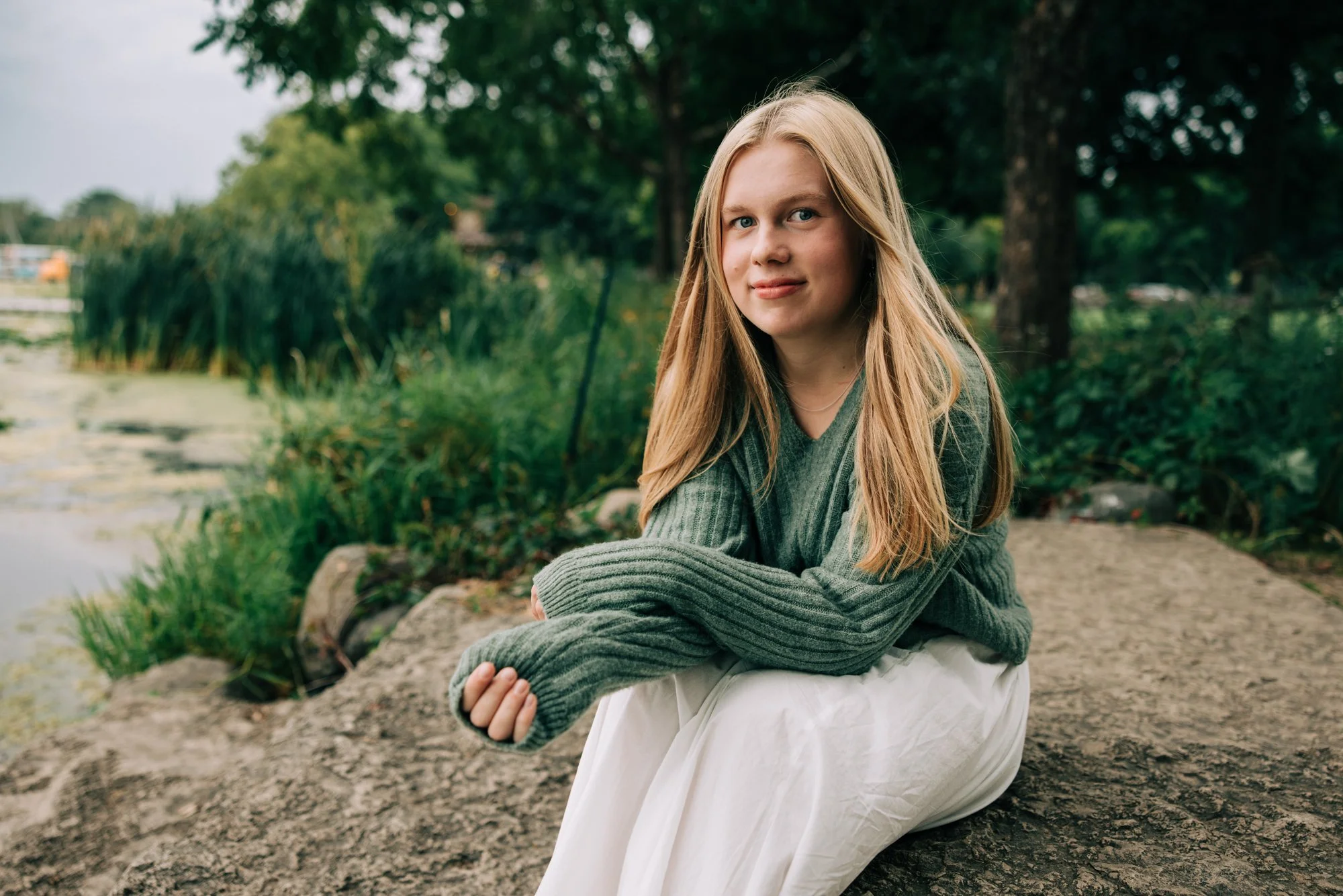 A young woman with blonde hair sitting on a rock by a lake, wearing a green sweater and white pants, with greenery and trees in the background.