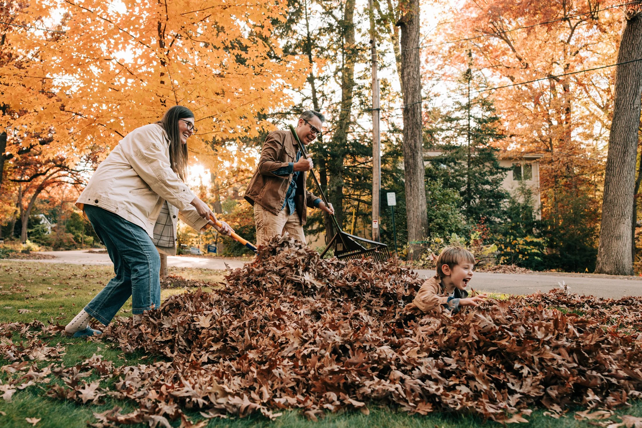 Family of three, including a woman, man, and young child, playing in a pile of fallen autumn leaves in a park with orange and yellow trees in the background.