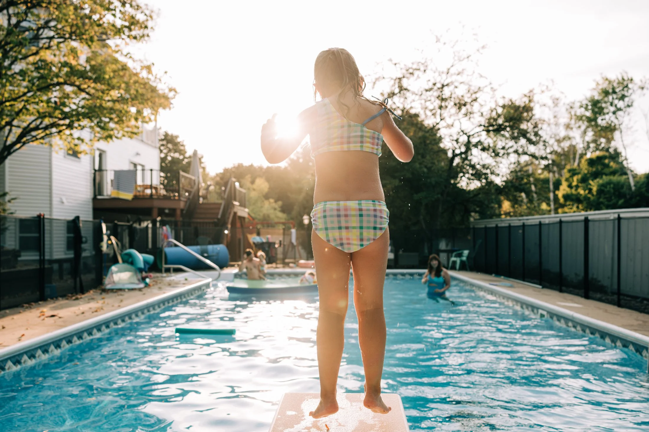 Young girl jumping into a backyard swimming pool during late afternoon, with other children swimming and water toys visible, surrounded by trees and houses.