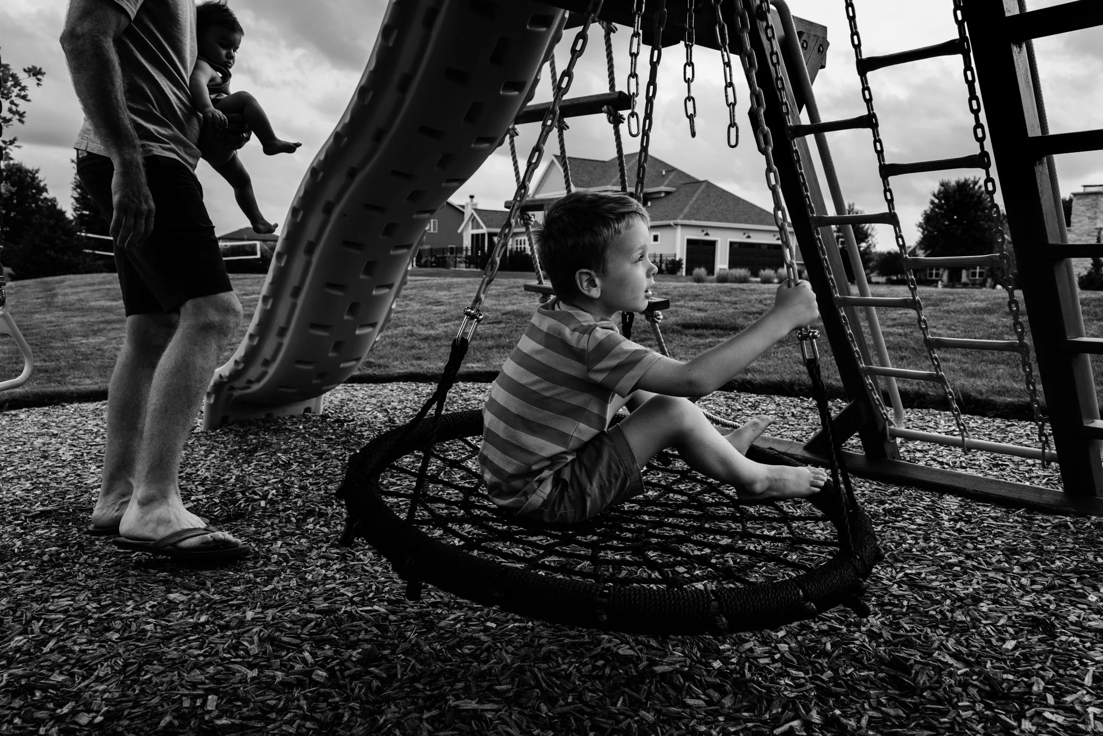 A young boy sitting on a circular rope swing at a playground, with an adult holding a small child nearby, all in a black and white outdoor setting.