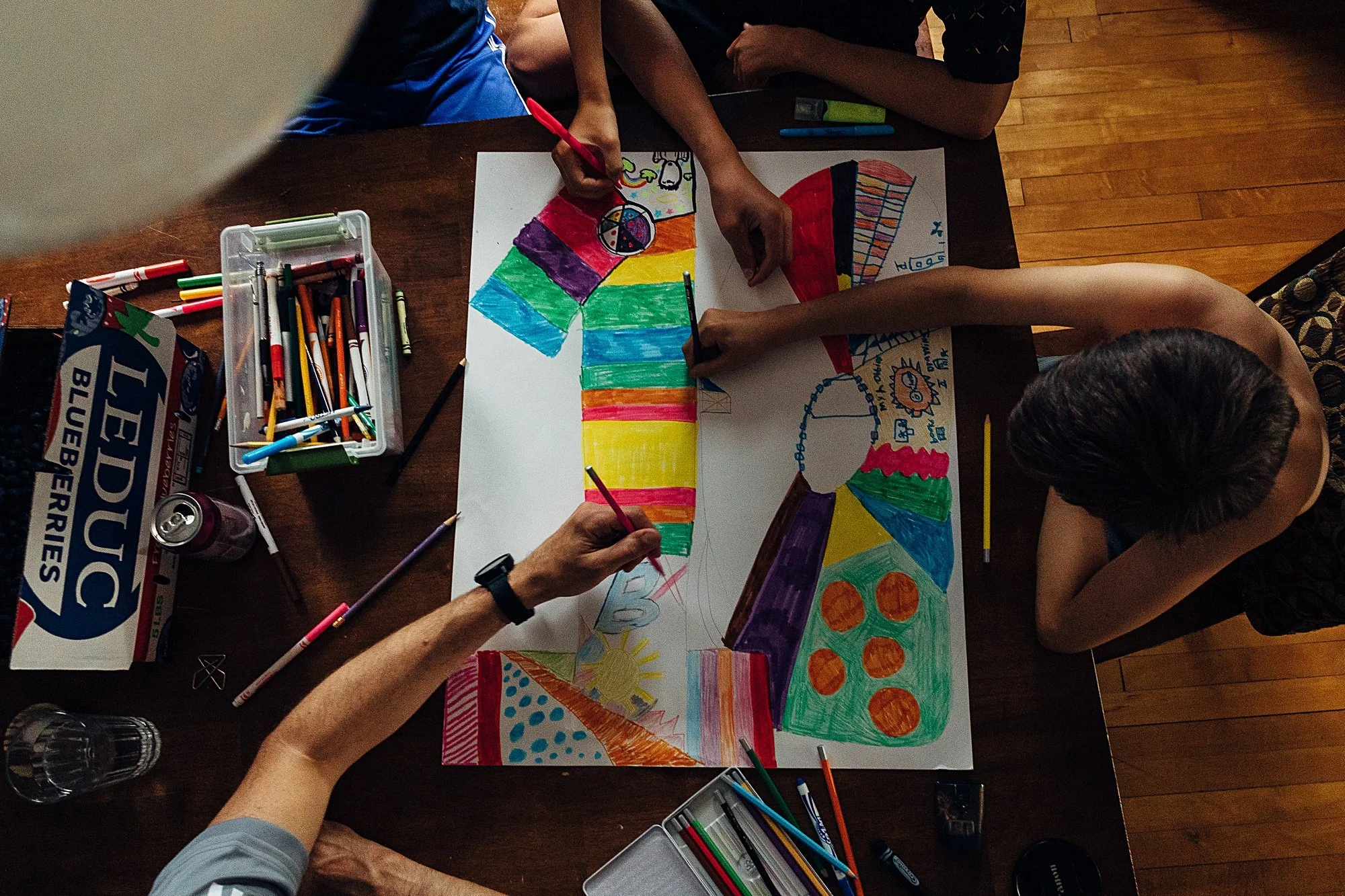 Children and adult coloring large poster with bright, colorful sections on a wooden table, surrounded by markers, pens, and a juice can.