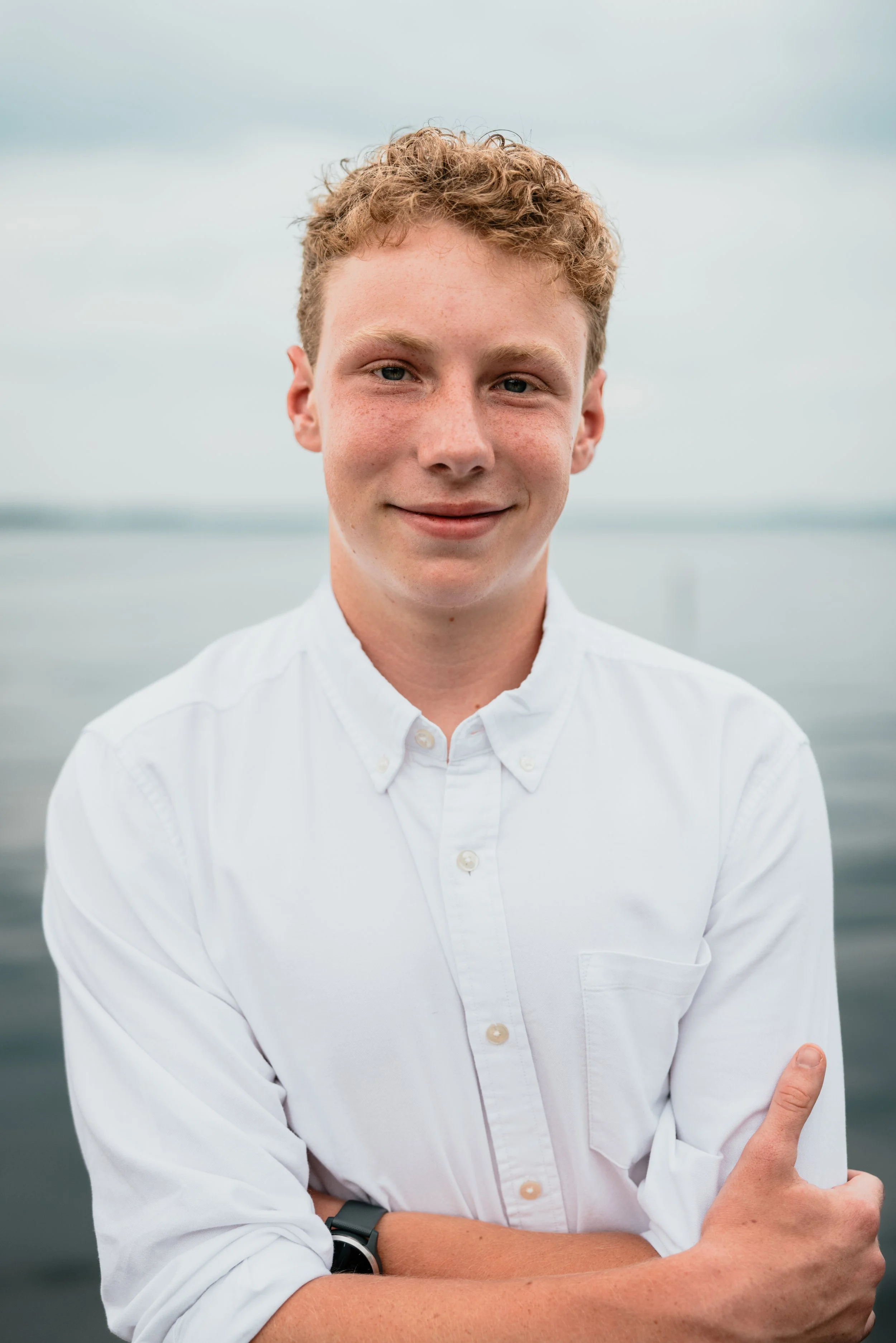 A young man with curly red hair and freckles, wearing a white button-down shirt, standing outdoors by a body of water with a cloudy sky in the background.