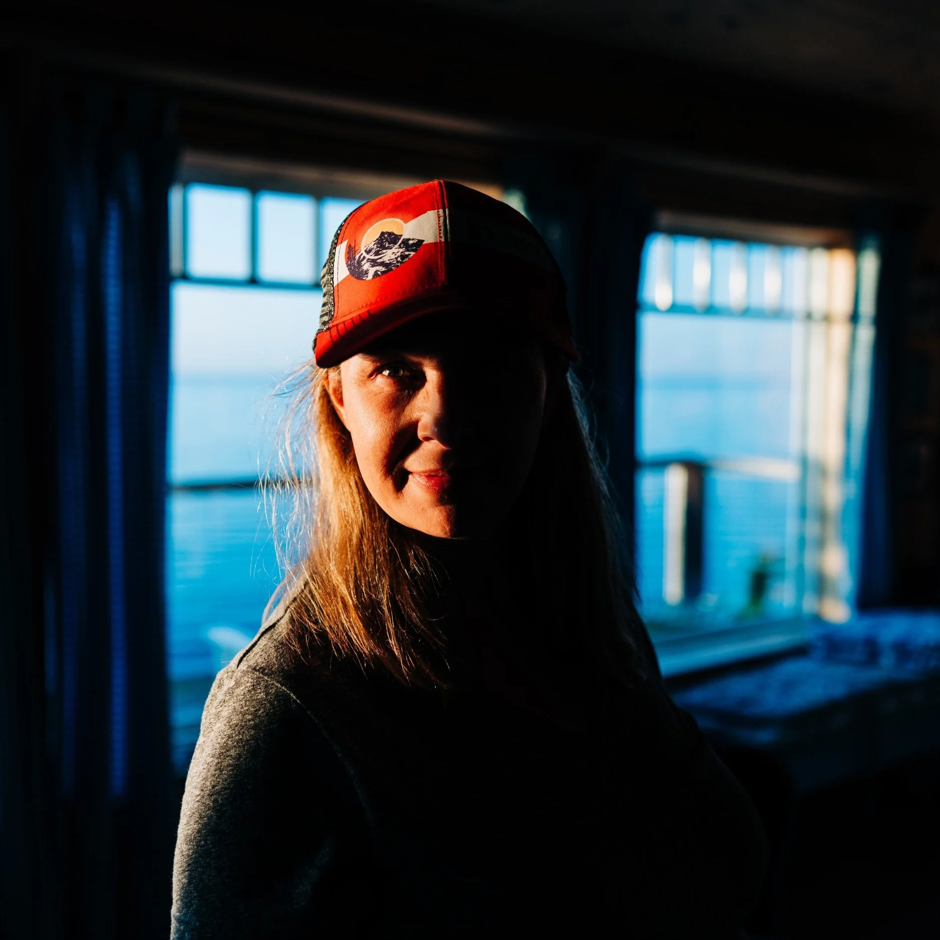 Photographer Jen Lucas wearing a red cap with a mountain logo, standing inside near windows overlooking water, with backlit sunlight creating shadows on her face.