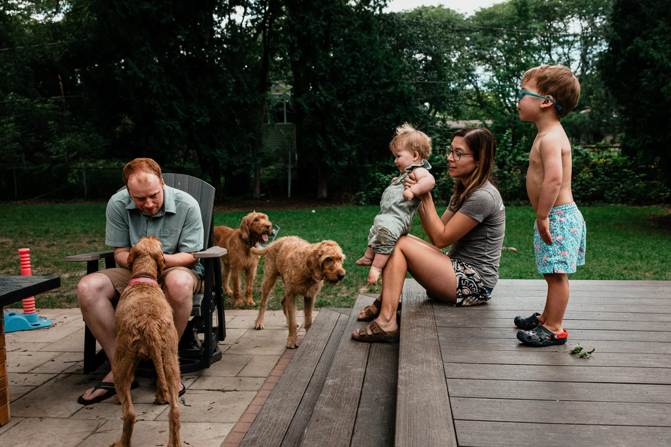 A family with two children and three dogs on a backyard patio with trees in the background.