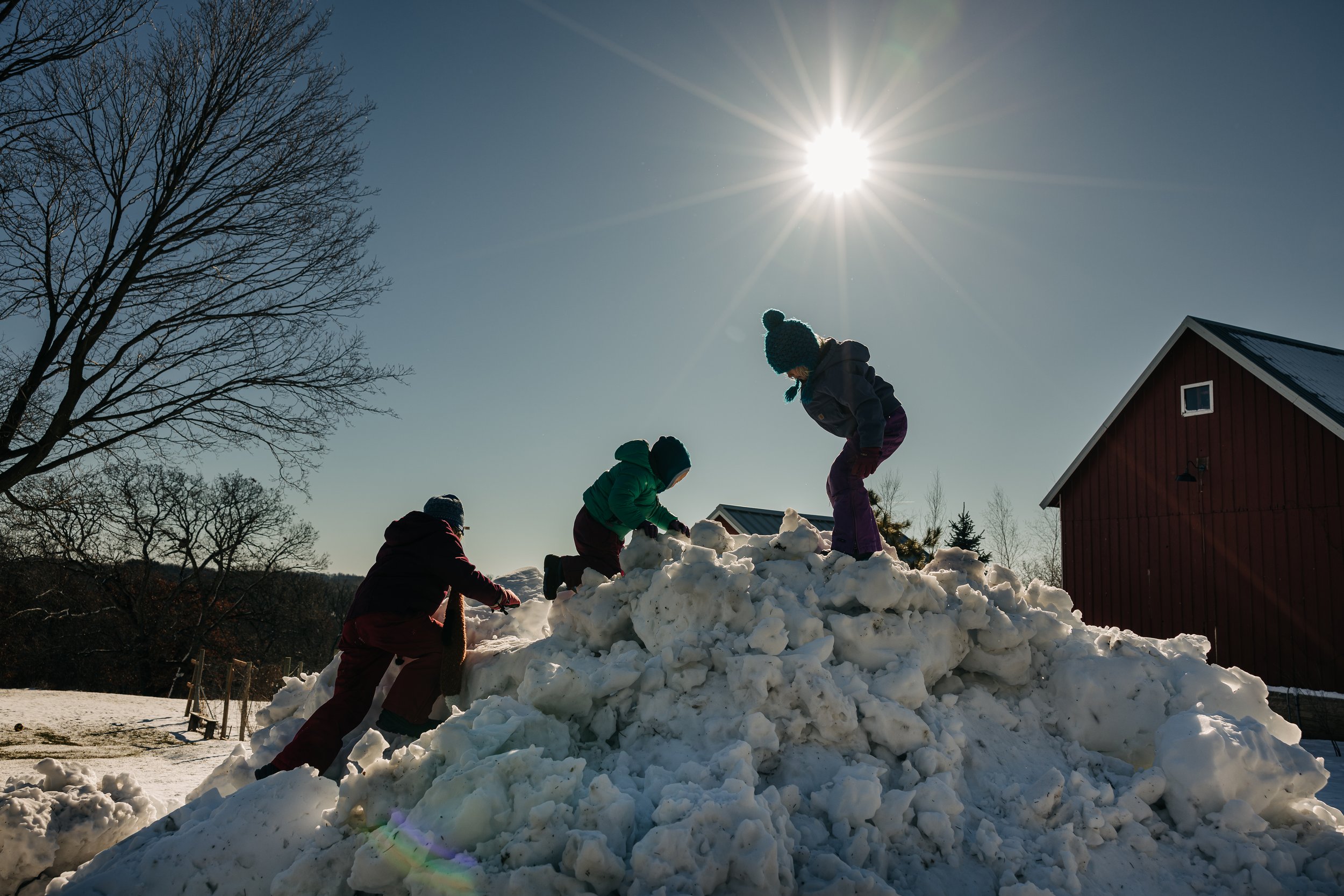 Children playing on snow pile outdoors under bright sun with clear sky, trees, and a red building in background.