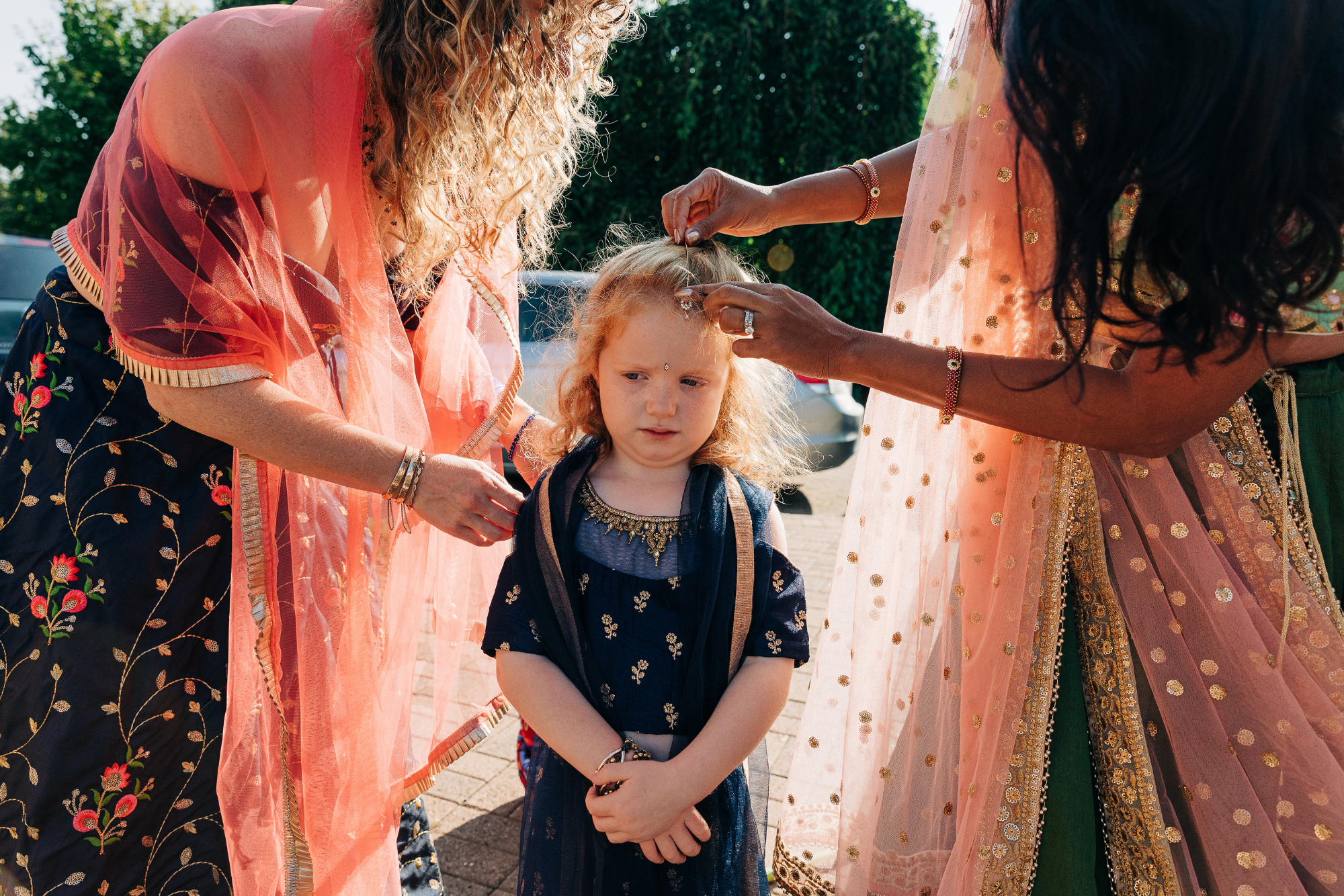 Two women decorating a young girl's forehead with traditional Indian accessories during a cultural event outdoors.