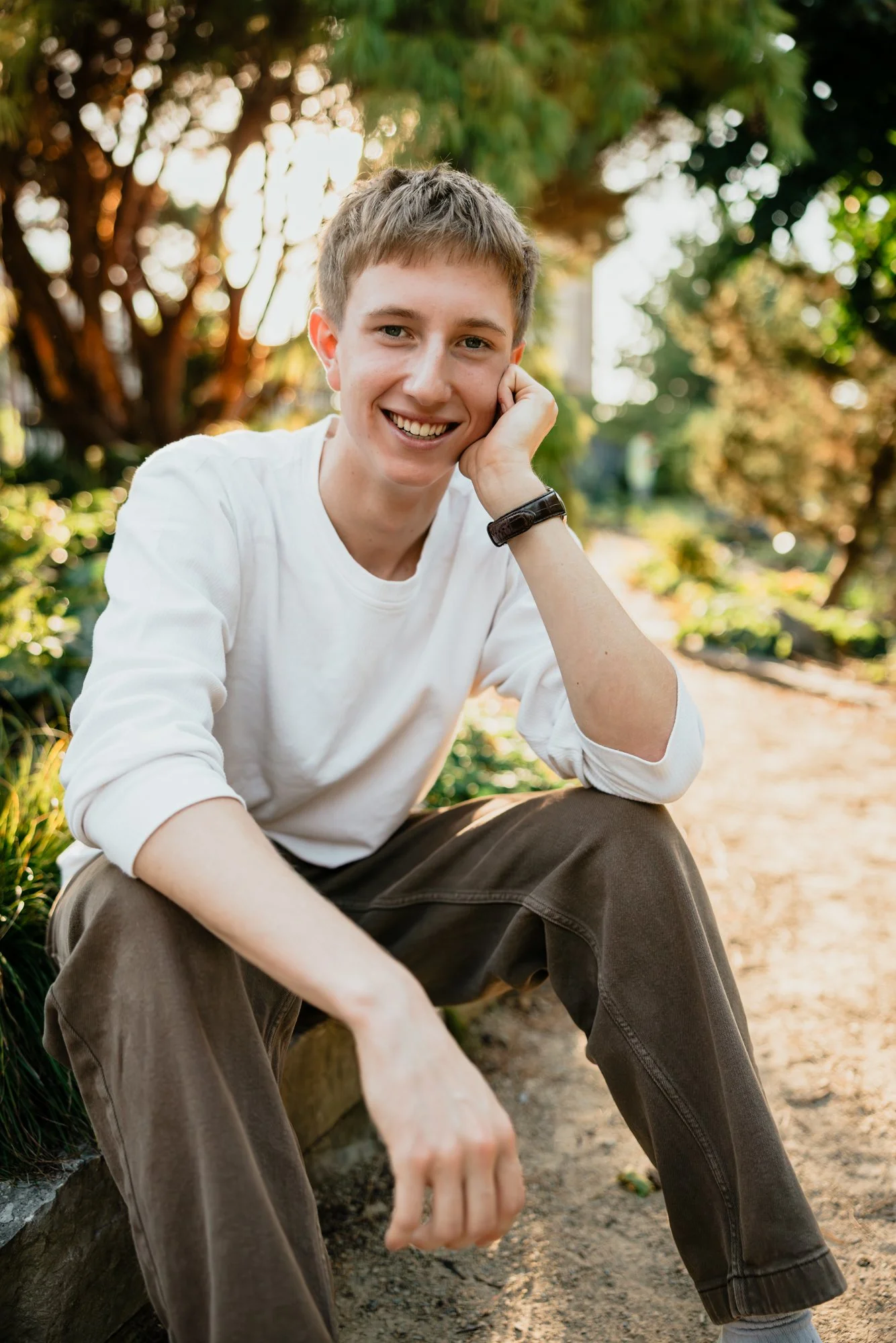 A young man with short hair, wearing a white long-sleeve shirt and brown pants, smiling while sitting outdoors on a dirt path surrounded by greenery and trees.