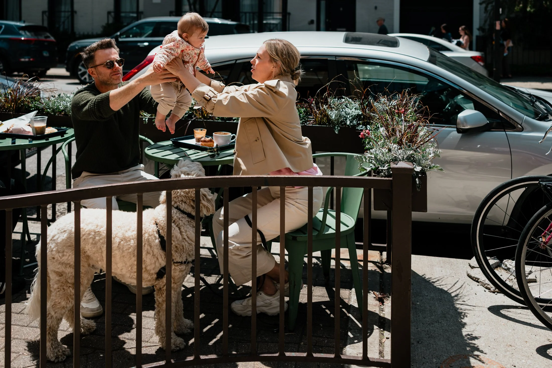 A man and woman are outside at a cafe with a child and a large fluffy dog. The man is holding the child, who is reaching toward the woman. The dog is on a leash and standing next to the table with food and drinks.
