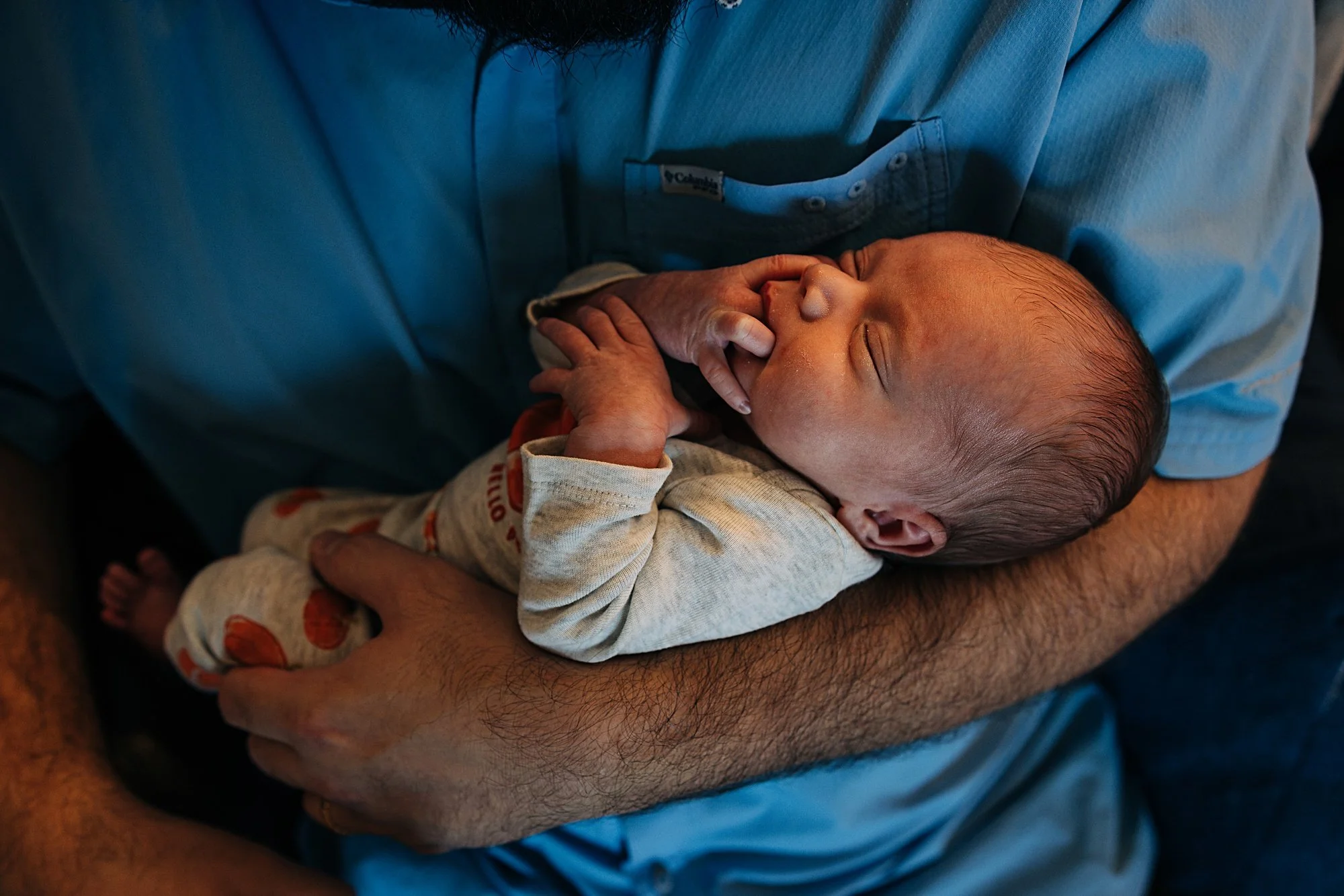 A man wearing a blue shirt gently holding a sleeping newborn baby in his arms.