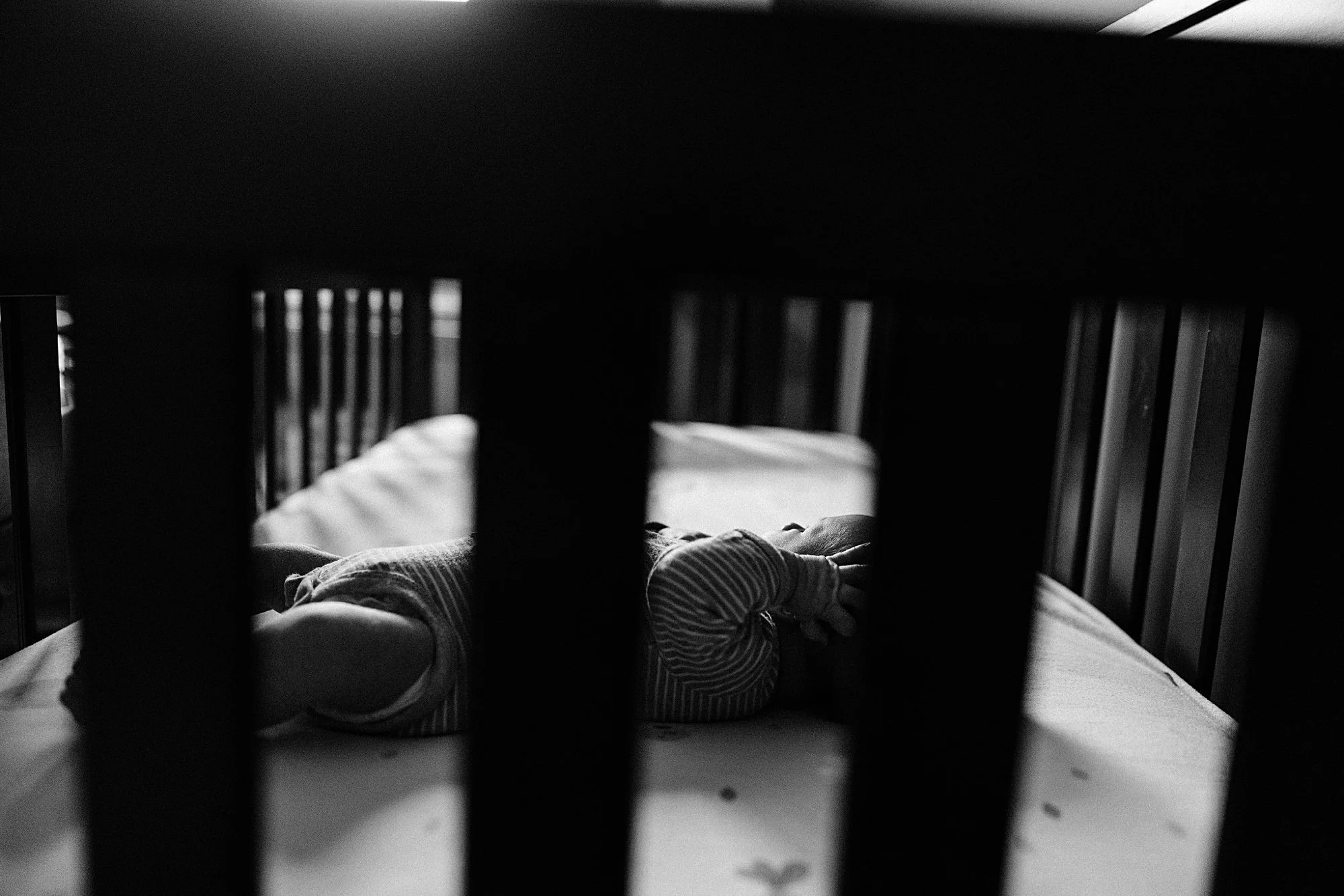 A baby lying in a crib, seen through the slats of the crib in black and white.