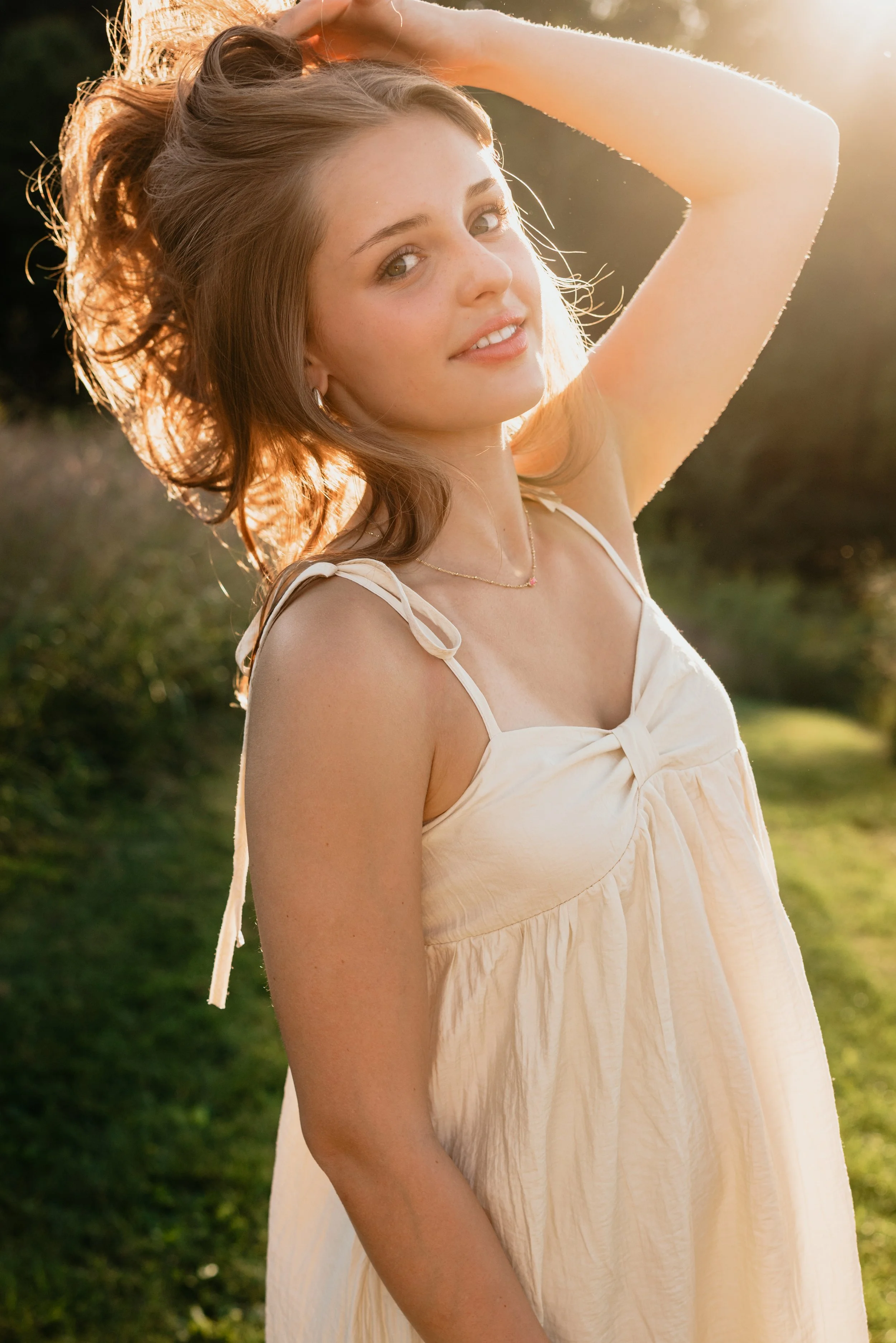 A young woman with light brown hair and blue eyes smiling outdoors at sunset, wearing a light-colored sundress with thin straps, with one hand raised touching her hair, in a grassy area with trees in the background.