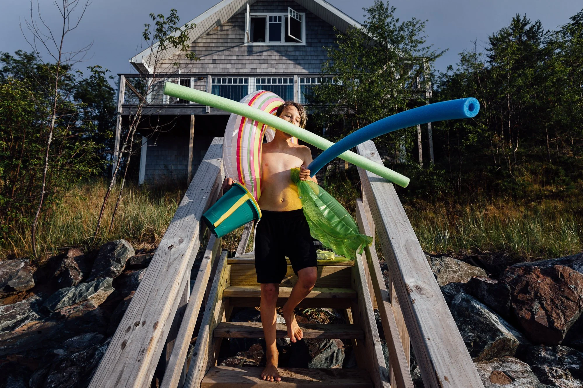 A young person with wet hair and beach attire carrying pool toys and a green float while descending a wooden dock staircase near the rocks and grass, with a house in the background.
