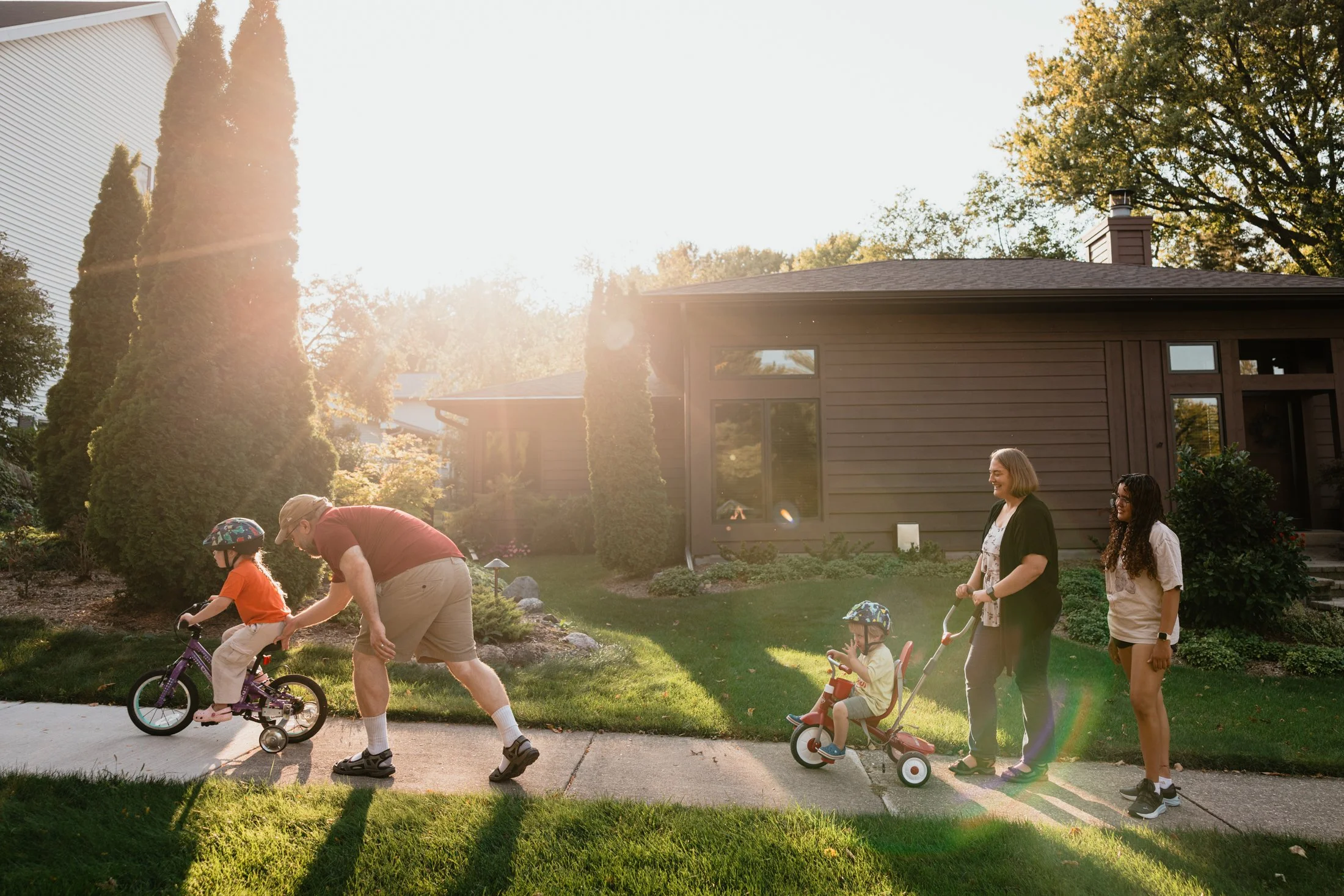 A family enjoying outdoor time on a sidewalk in front of a house during sunset. A man helps a young girl ride a bicycle with training wheels, a woman pulls a child's tricycle, and another woman walks beside a teenager. The scene is sunny with a well-