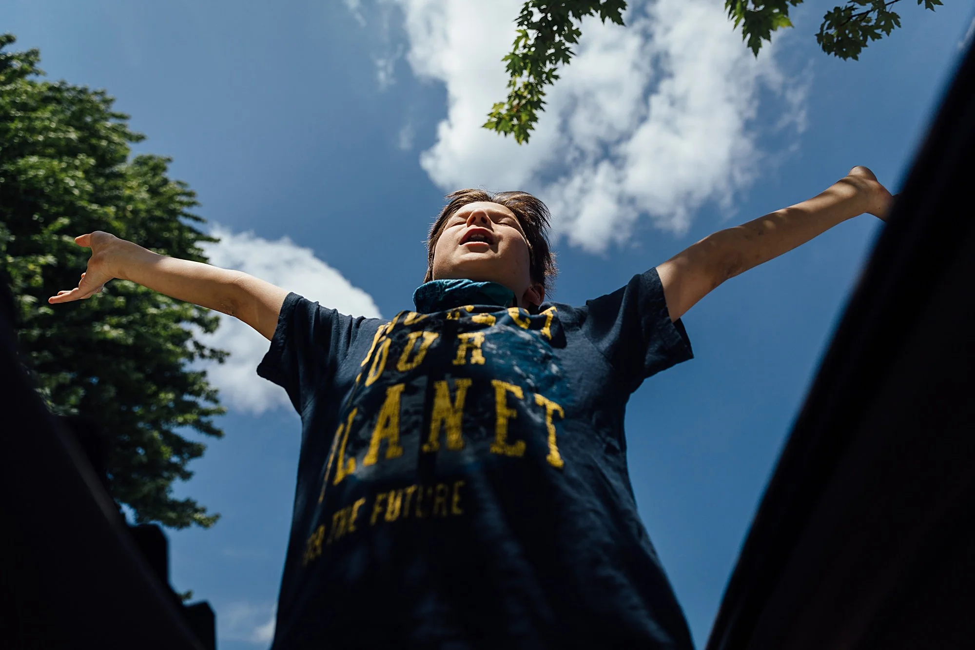 A young boy with arms outstretched, looking upward, standing outdoors against a blue sky with white clouds and green trees, wearing a black t-shirt with yellow lettering.