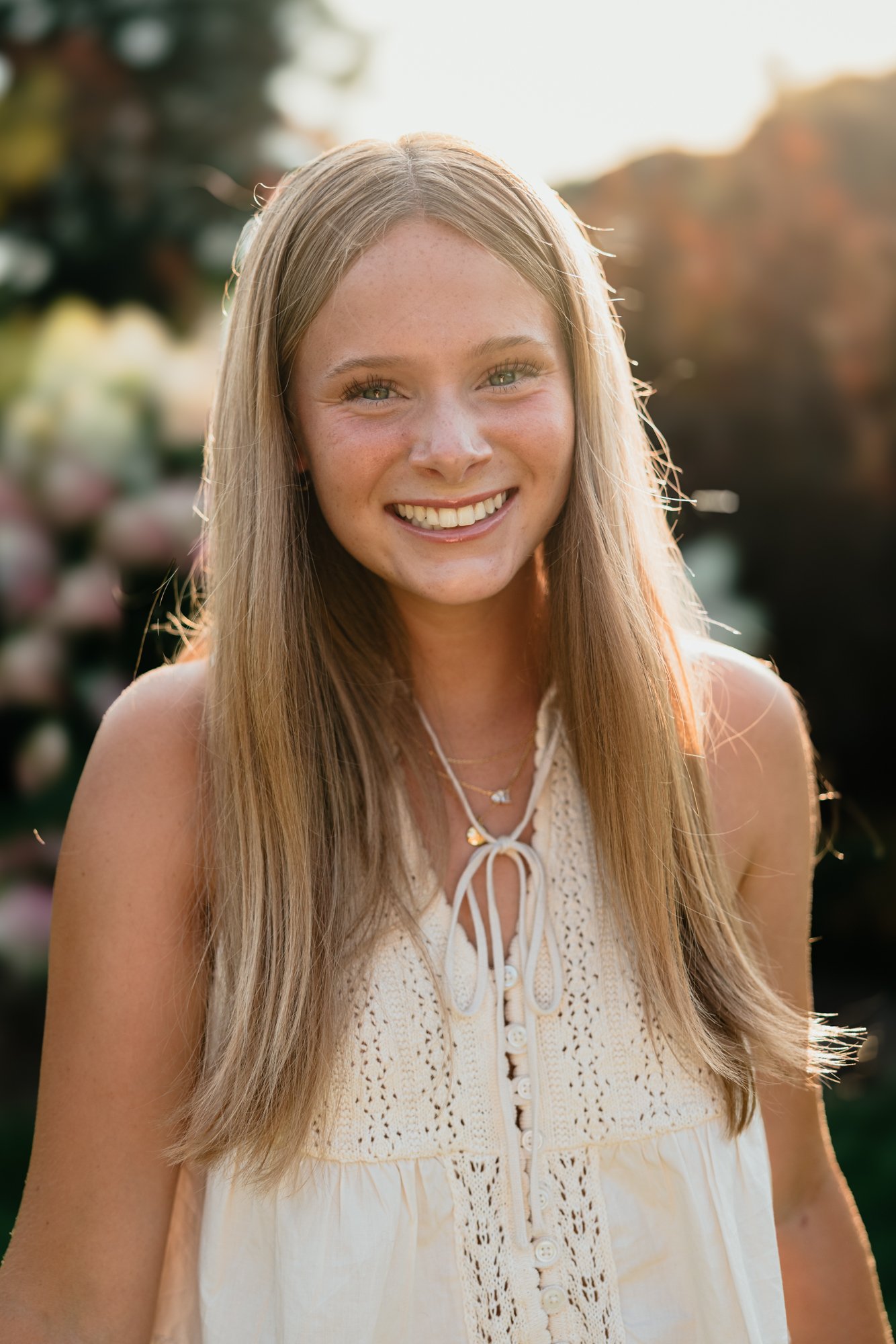 A young woman with long blonde hair smiling outdoors during golden hour. She is wearing a cream-colored crochet top and layered necklaces.
