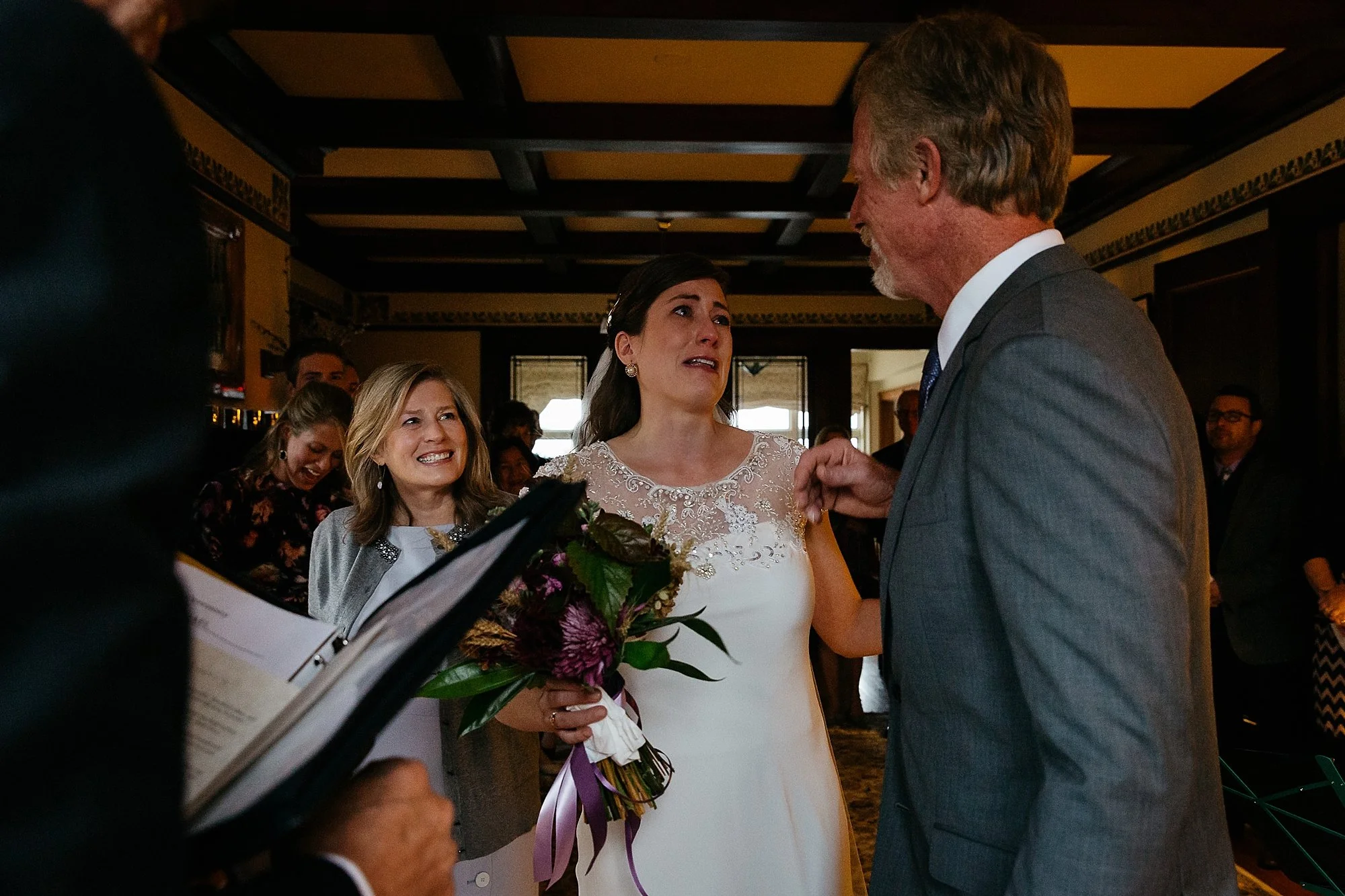 A woman in a white wedding dress appears emotional while speaking to a man in a gray suit at a wedding ceremony. A woman holding a bouquet of flowers smiles nearby, with several guests in the background.