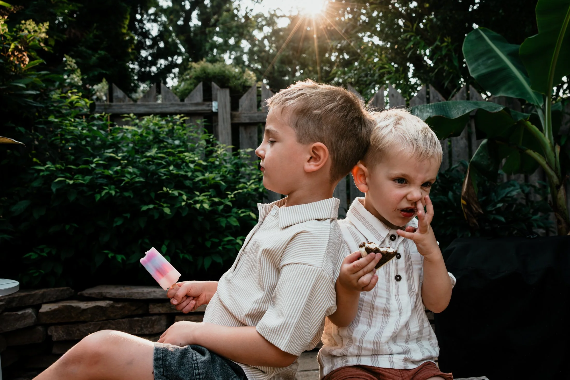 Two young boys sitting back-to-back outdoors, one eating a slice of cake and the other holding an ice cream bar, with a wooden fence and greenery in the background during sunset.