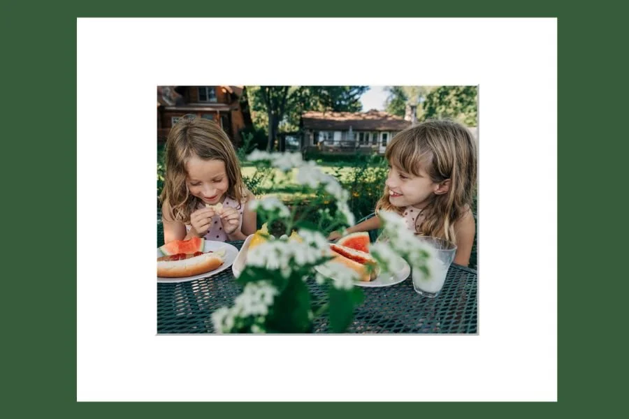 Two young girls are sitting at an outdoor table with watermelon slices and a drink, smiling and enjoying each other's company with a house and trees in the background.
