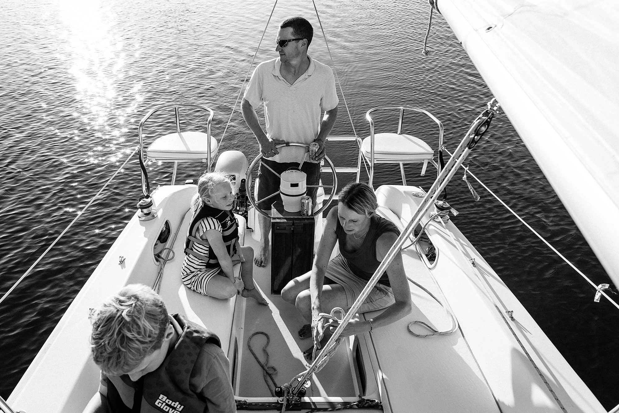 Family on a sailboat enjoying a sunny day, with some members sitting and others standing, surrounded by water.