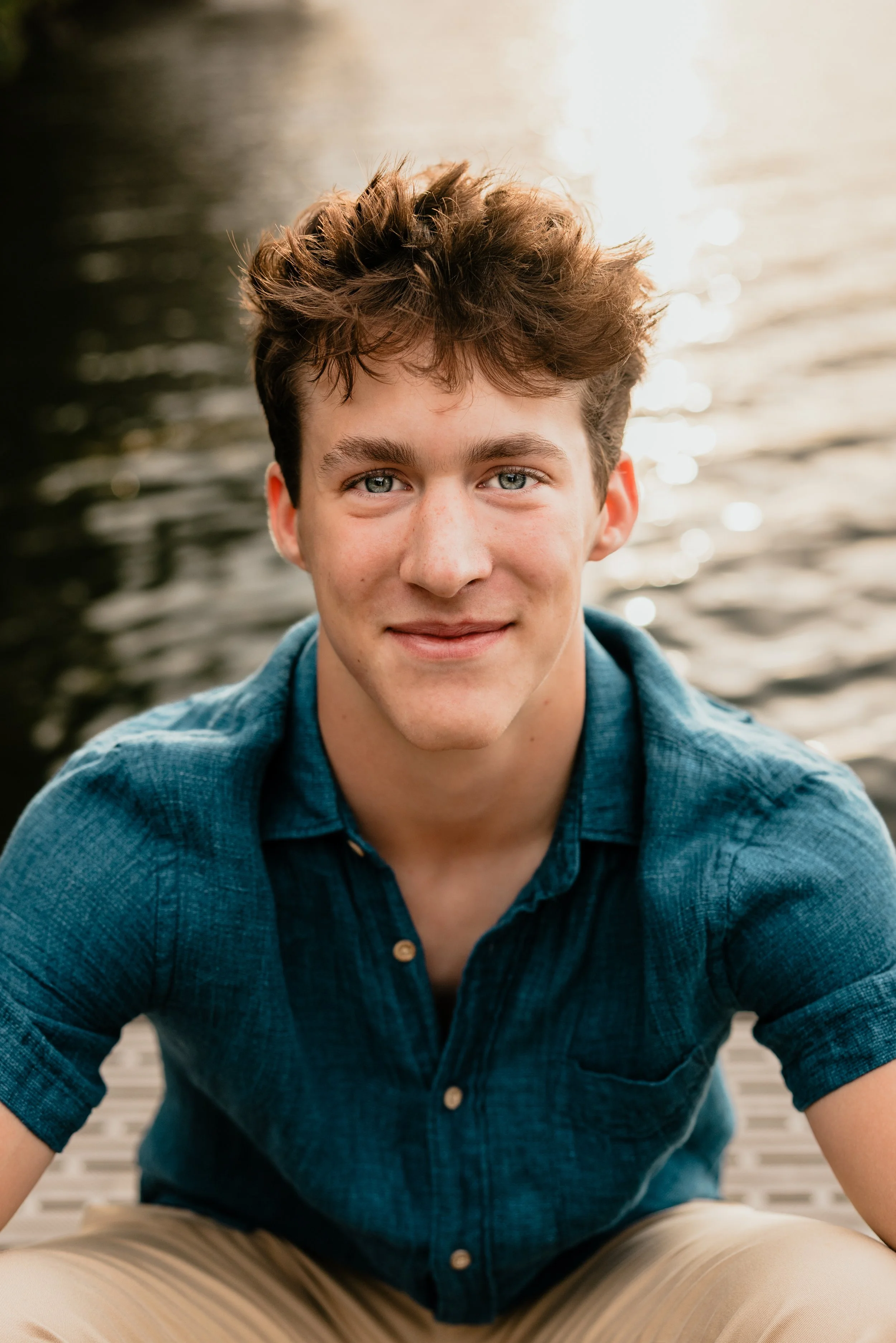 A young man with curly brown hair and blue eyes smiling while sitting near a body of water during sunset, wearing a blue short-sleeve button-up shirt.