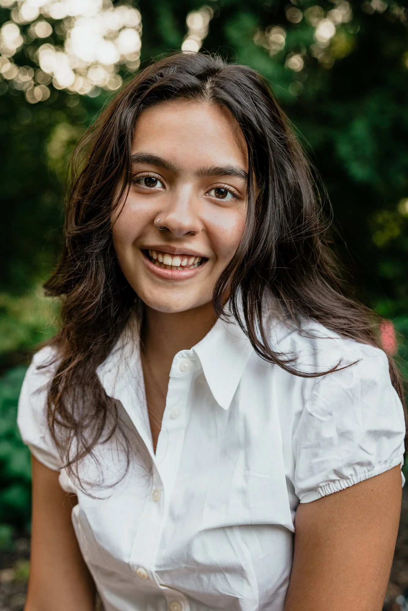 A young woman with dark brown, wavy hair, wearing a white short-sleeved button-up shirt, smiling outdoors with a blurred green natural background.