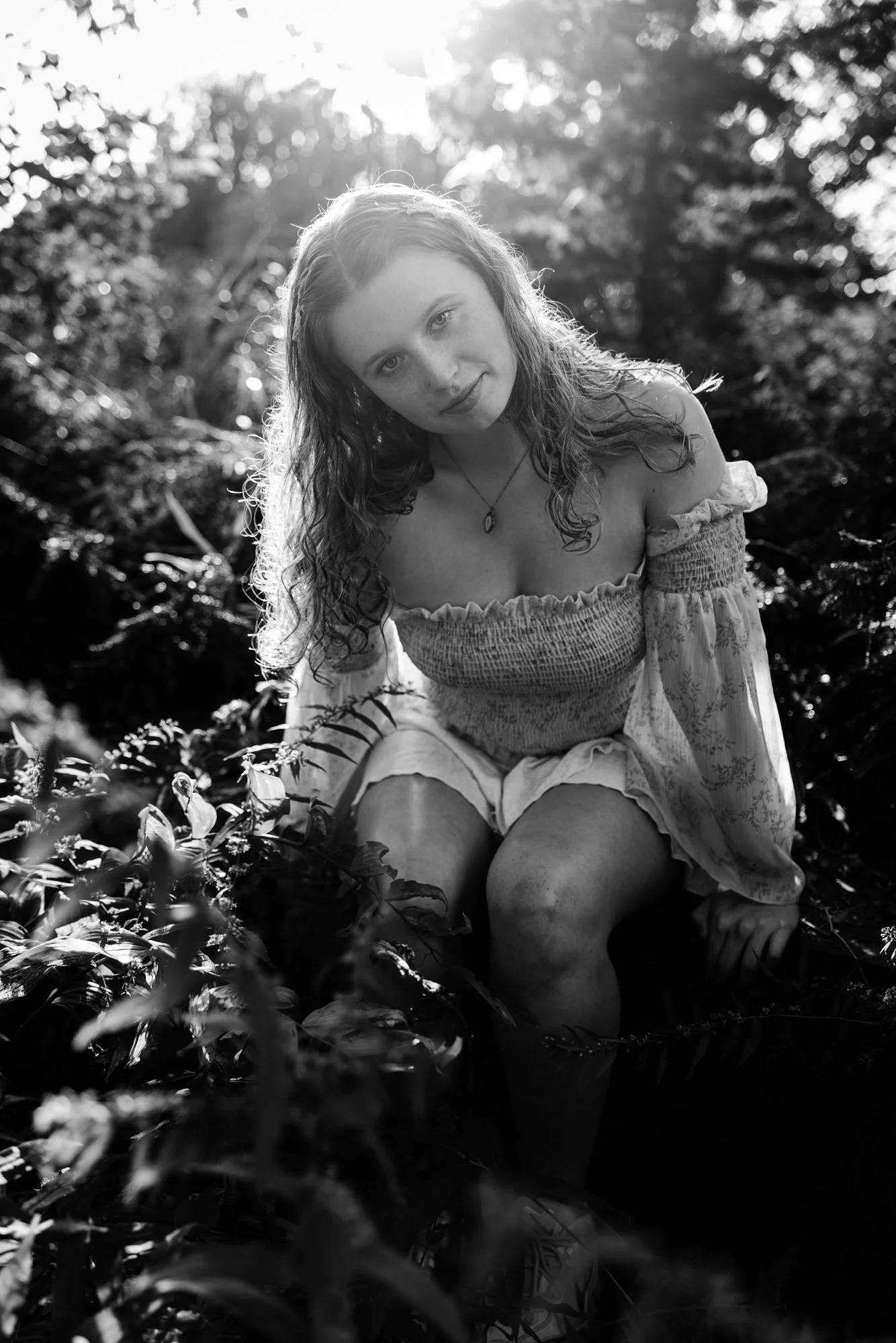 A young woman with wet, curly hair sitting outdoors amid foliage, looking into the camera, sunlight behind her, in black and white.