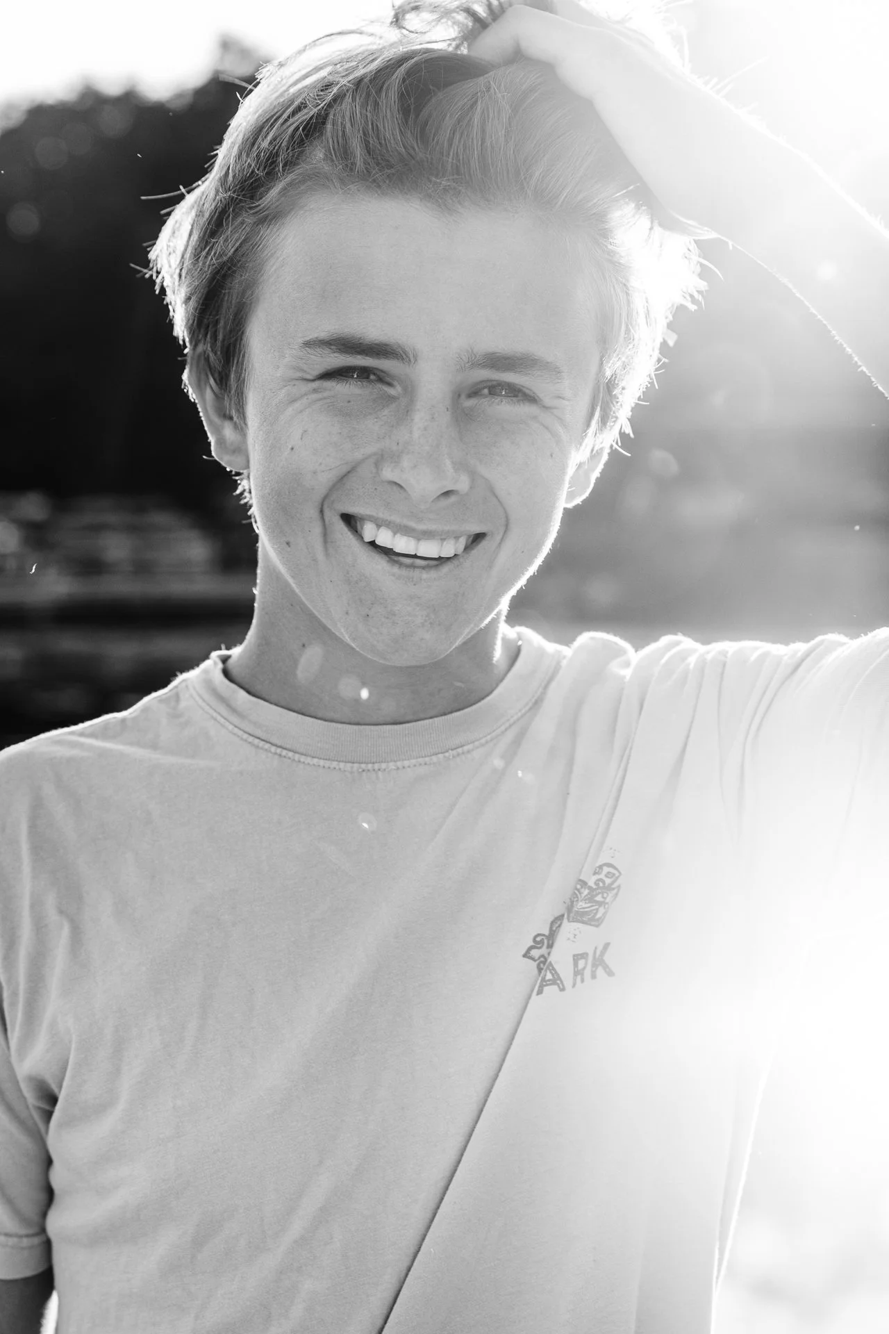 A young man smiling outdoors in sunlight, wearing a light-colored t-shirt, with one hand on his head.