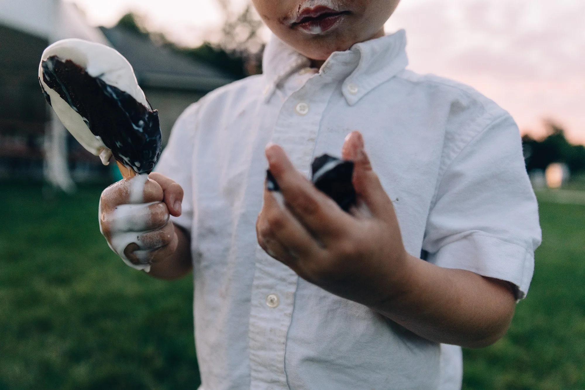 A young person holding a chocolate and vanilla ice cream popsicle with a bite taken out of it, wearing a white button-up shirt, outdoors during sunset.