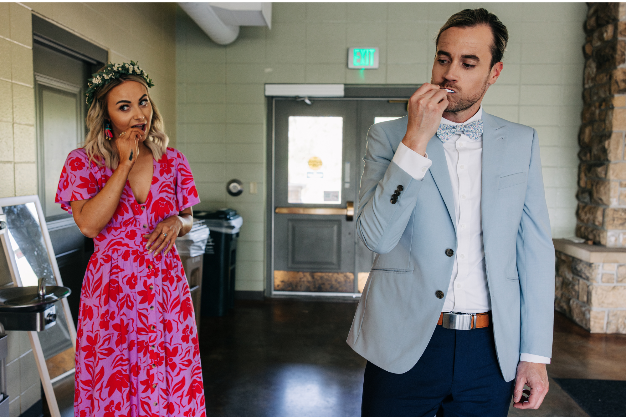 A man in a light blue suit and a woman in a pink floral dress are standing indoors, both touching their lips with fingers in a pose suggestive of worry or contemplation, with an exit door in the background.