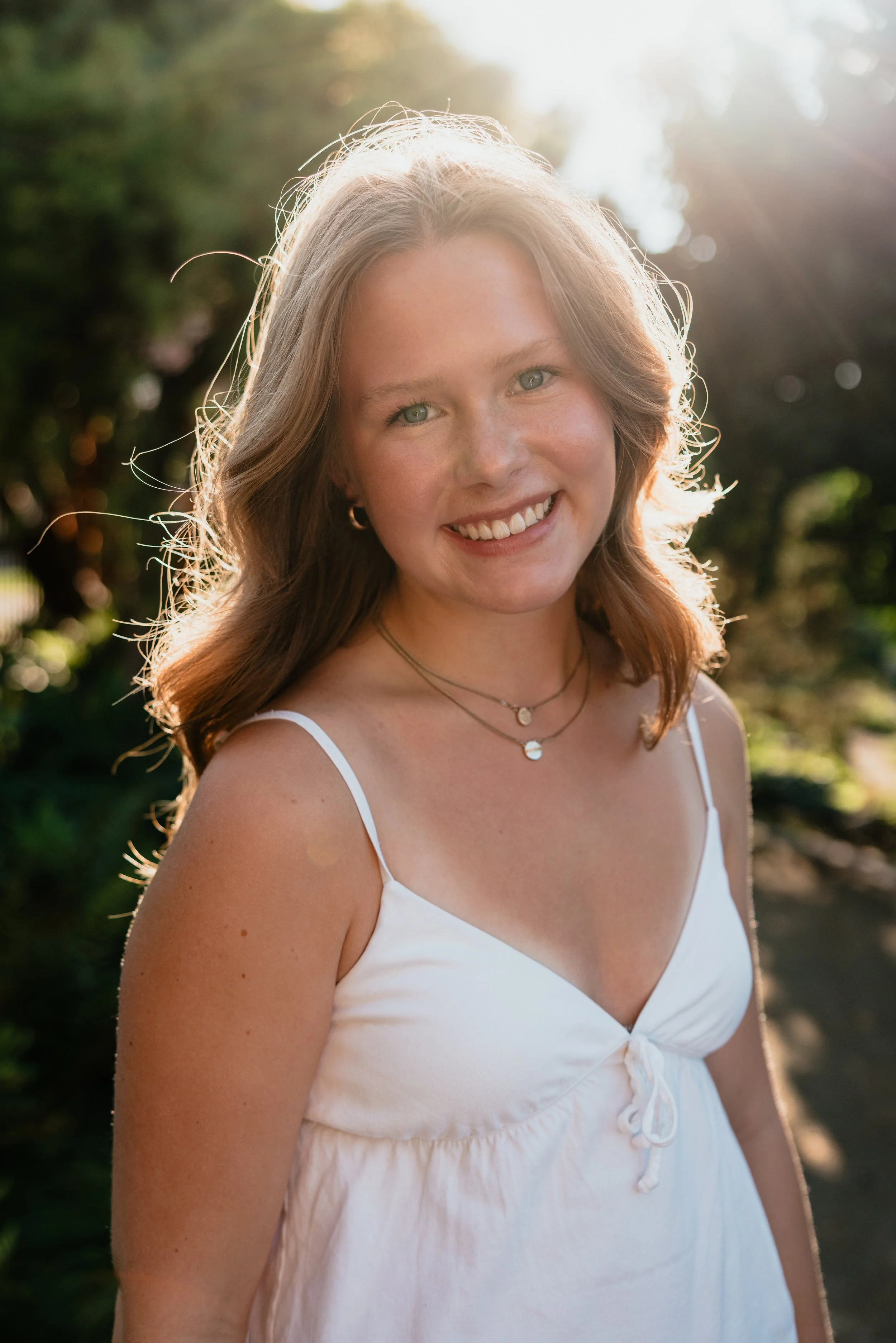 A young woman with wavy, shoulder-length hair, smiling outdoors in sunlight, wearing a white spaghetti strap dress and layered necklaces.