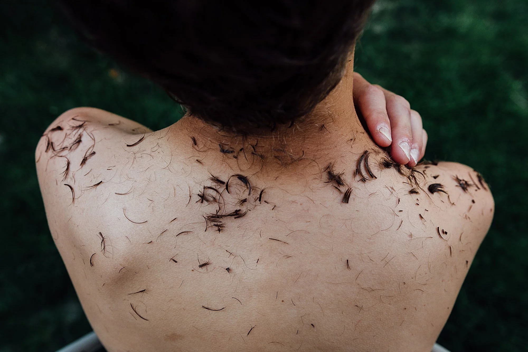 Close-up of a person's back and neck with shaved hair, some hair clippings on their skin, and someone gently touching their neck.