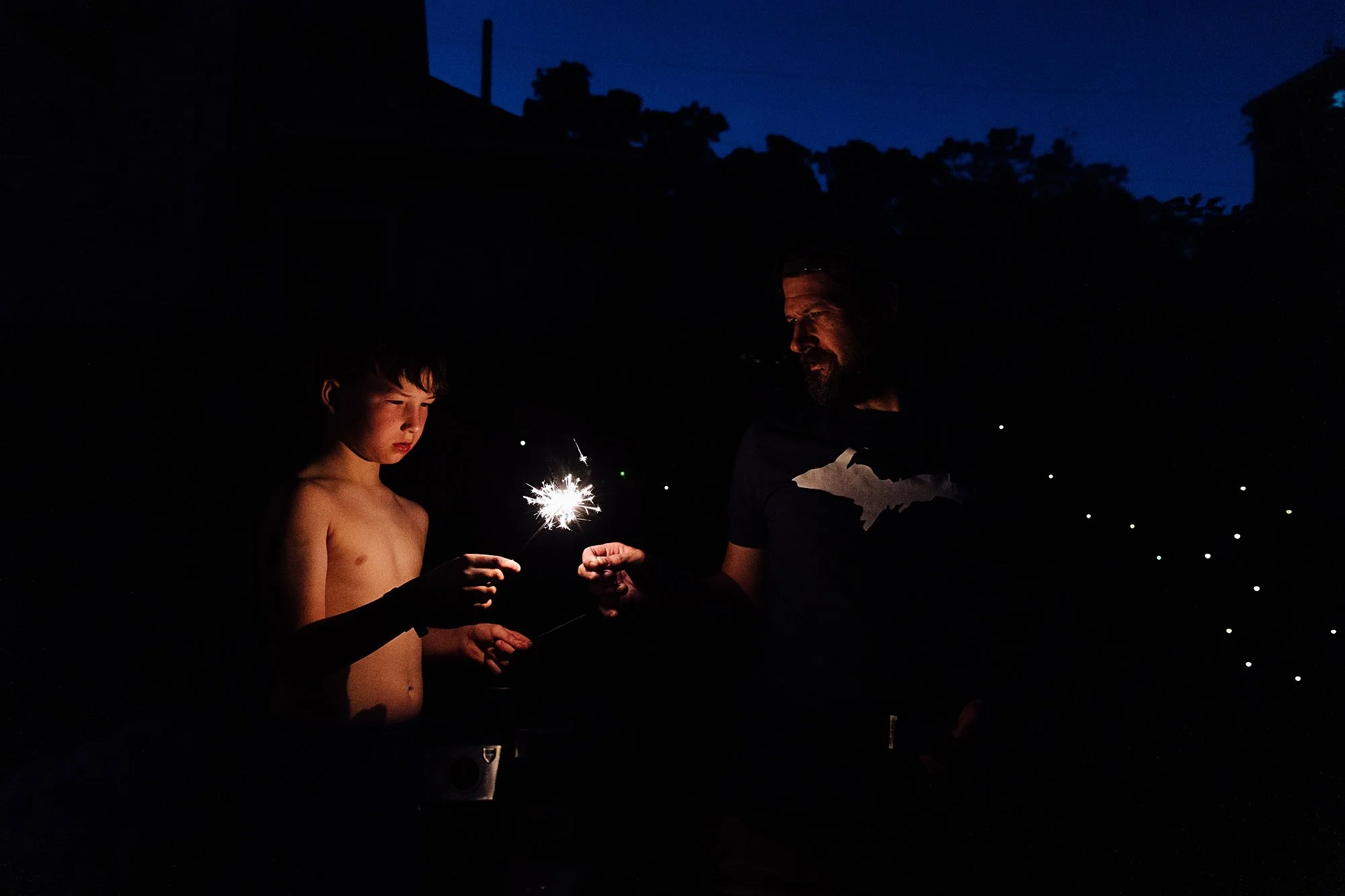 A young boy and an adult man are outdoors at night, holding sparklers with sparks flying in a dark setting. The background shows a night sky and silhouette of trees and buildings.