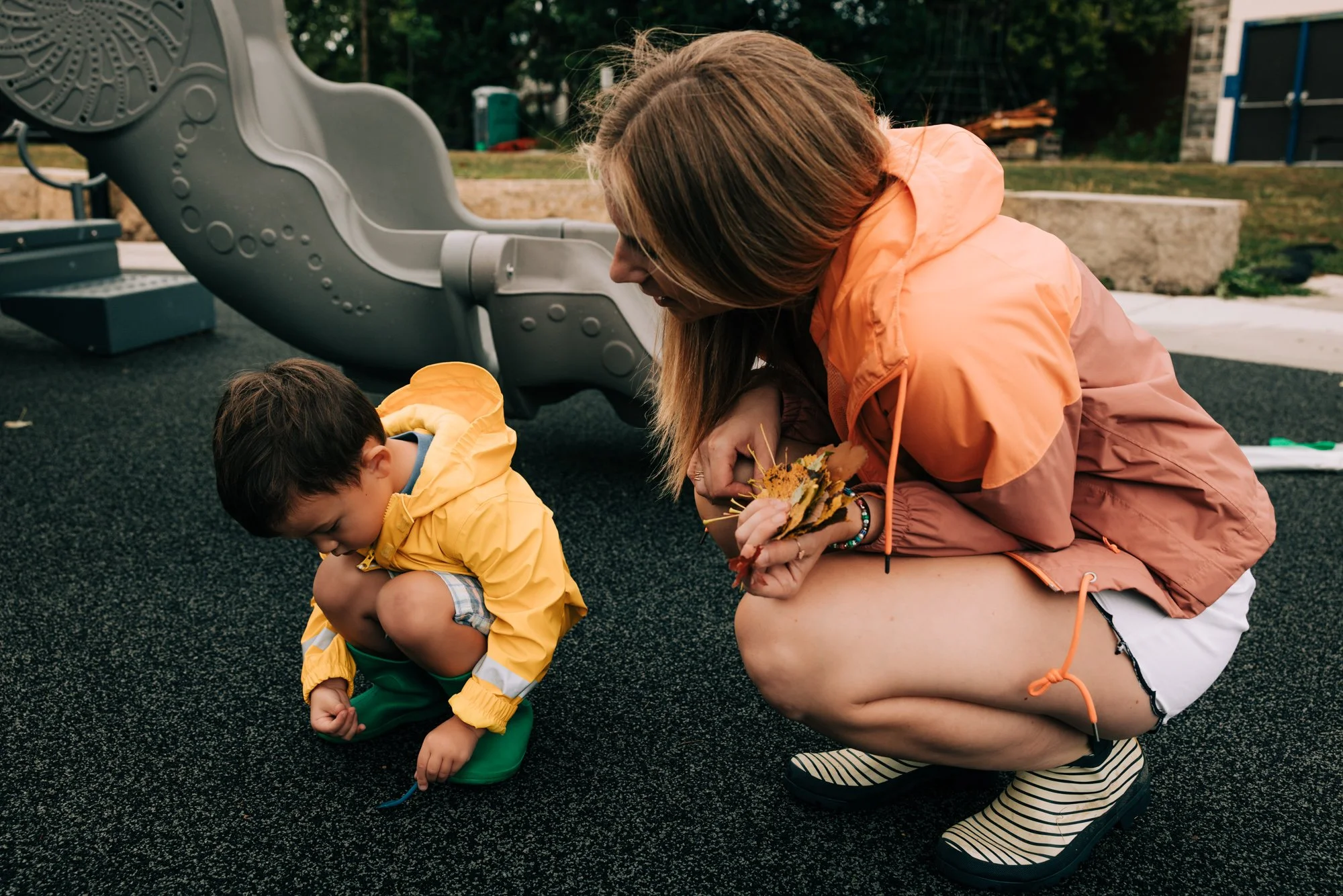 A woman and a young boy crouch down on a playground surface, with the woman holding a bunch of leaves and the boy looking at something on the ground.