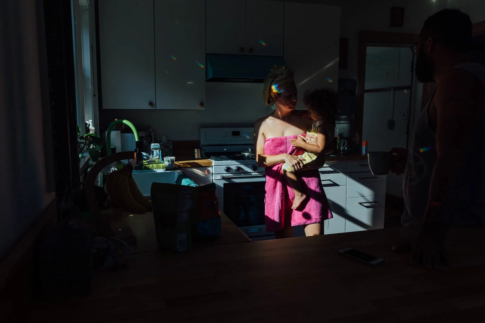 A woman holding a young girl in a kitchen with sunlight streaming in, casting a rainbow of light reflections on the walls and cabinets. The woman is wrapped in a pink towel, and the girl is wearing a yellow dress. An arm of a man holding a mug is visible on the right side of the image.