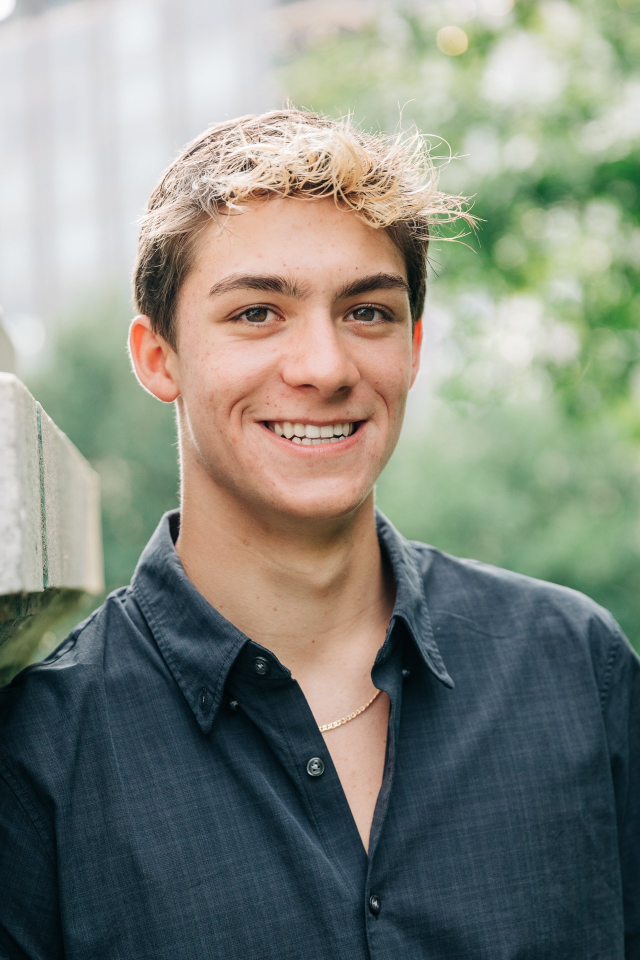 A young man with light brown, slightly curly hair smiling outdoors with a blurred green background.