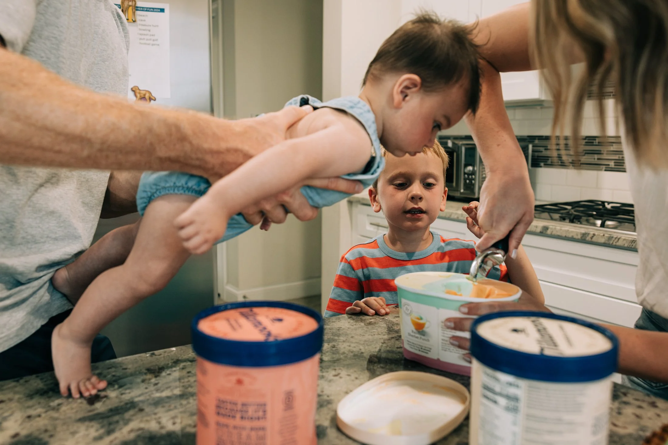 A young child is being helped to scoop ice cream from a tub into a bowl by an adult, while another child observes. The scene takes place in a kitchen with a countertop and several containers of ice cream in the foreground.