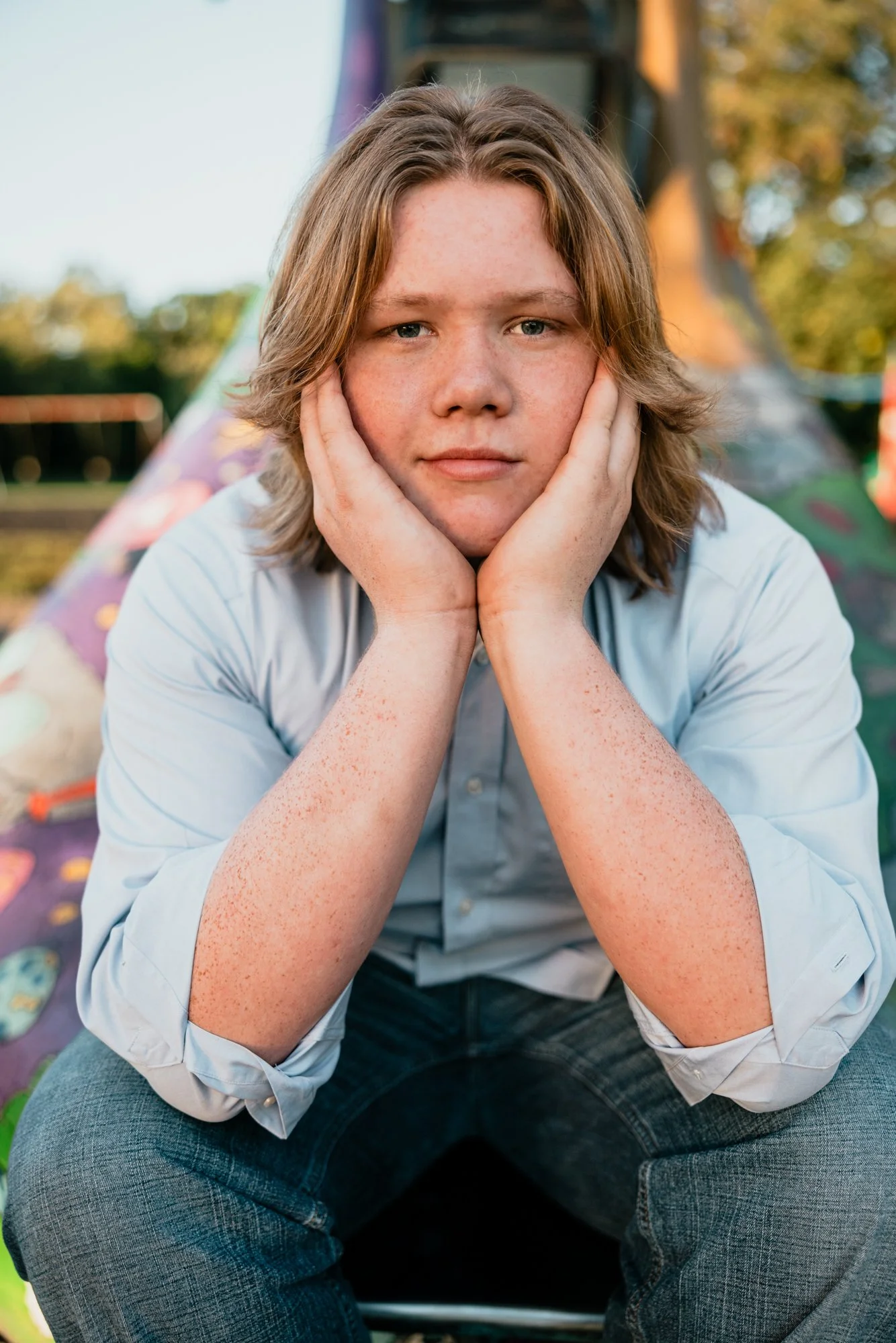 A teenage boy with light brown, wavy hair and light freckles, sitting outdoors with his chin resting on his hands, looking directly at the camera, with a colorful park or playground in the blurred background.
