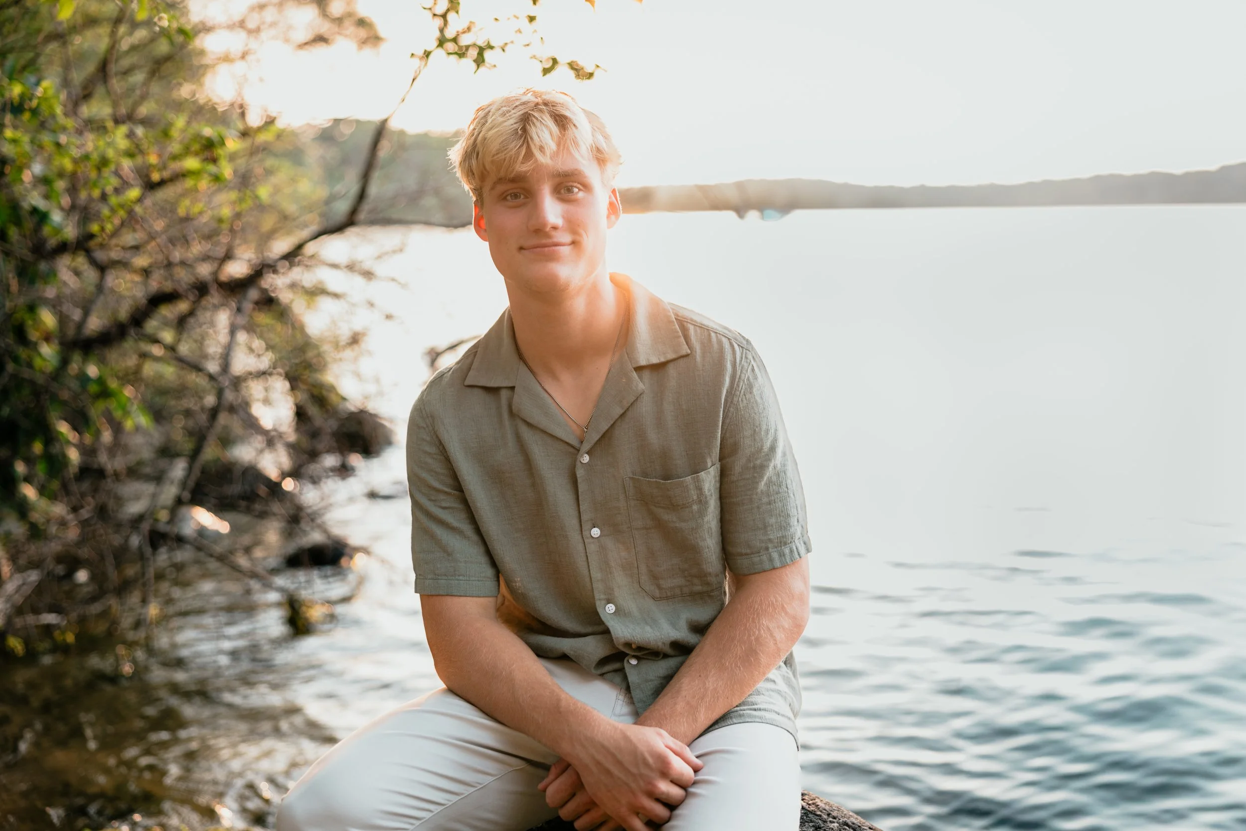 A young man with blonde hair sitting by a body of water during sunset, wearing a beige short-sleeved button-up shirt and light-colored pants.