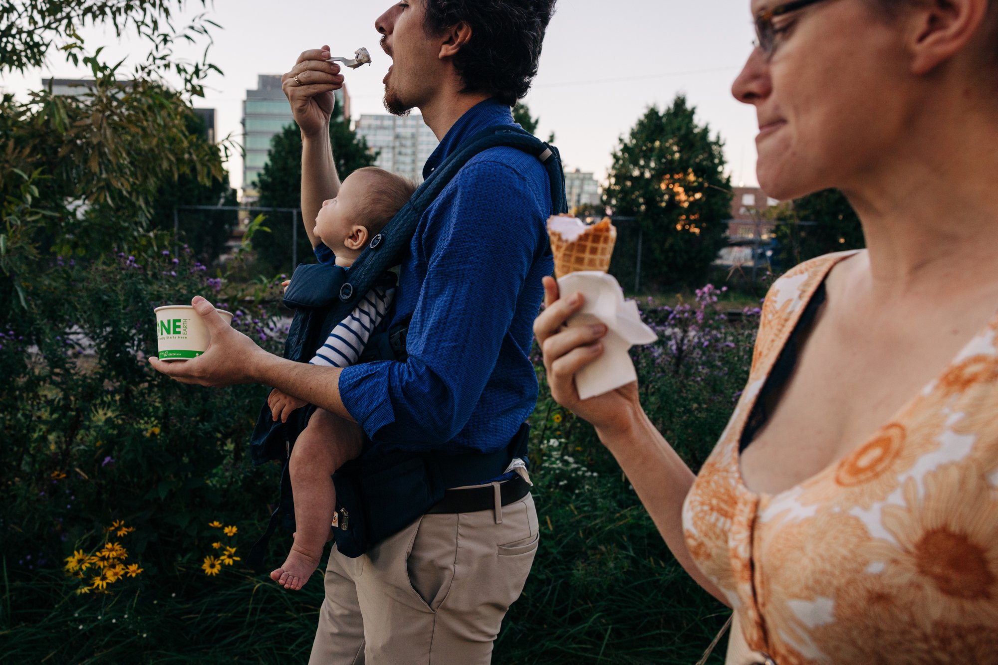 A man carrying a baby in a baby carrier eating ice cream while a woman standing next to them holds a cone of ice cream in her hand outdoors in the evening.