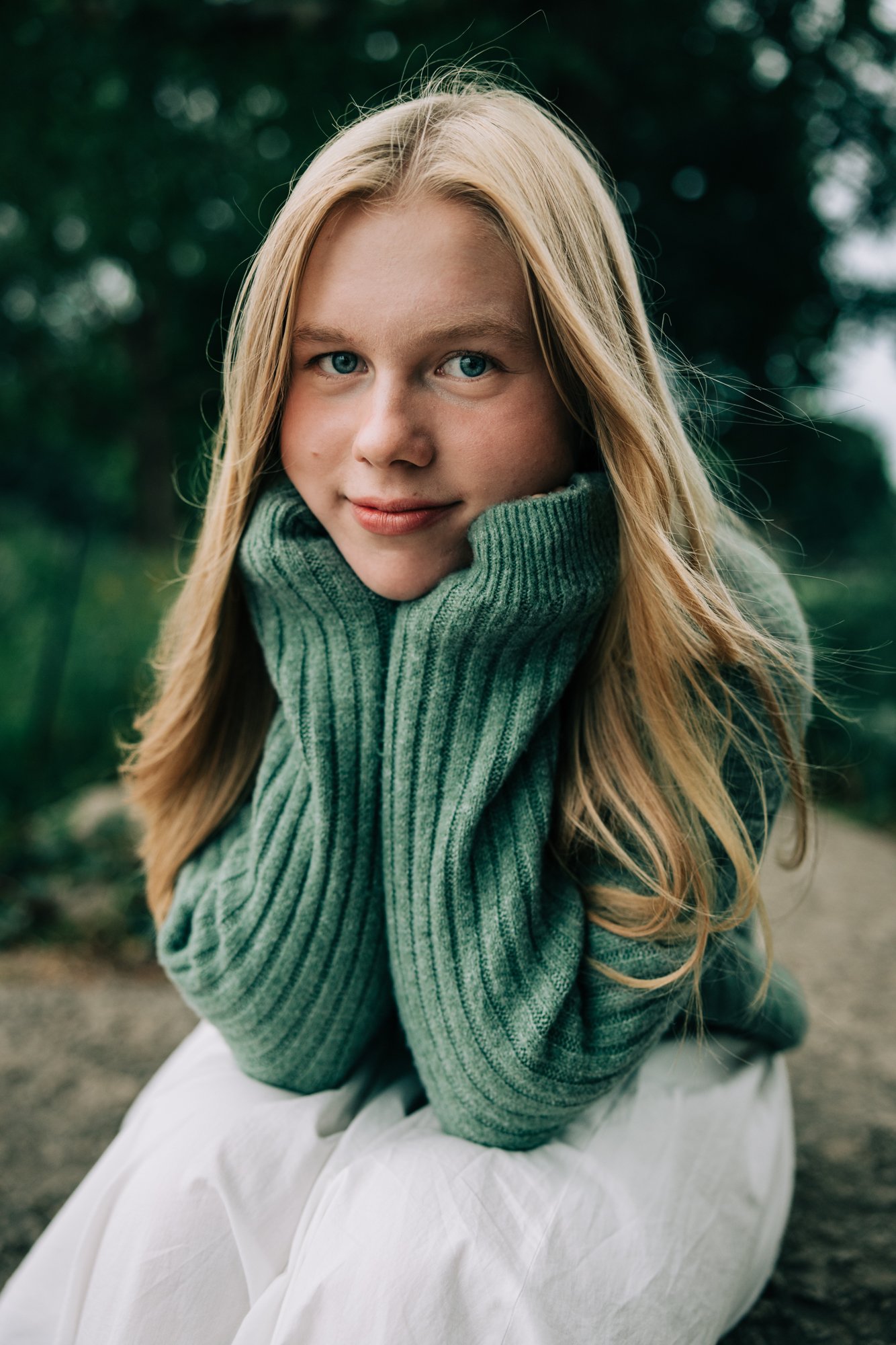 A young girl with long blonde hair and blue eyes, wearing a green knit sweater, sitting outdoors with green blurred trees in the background.