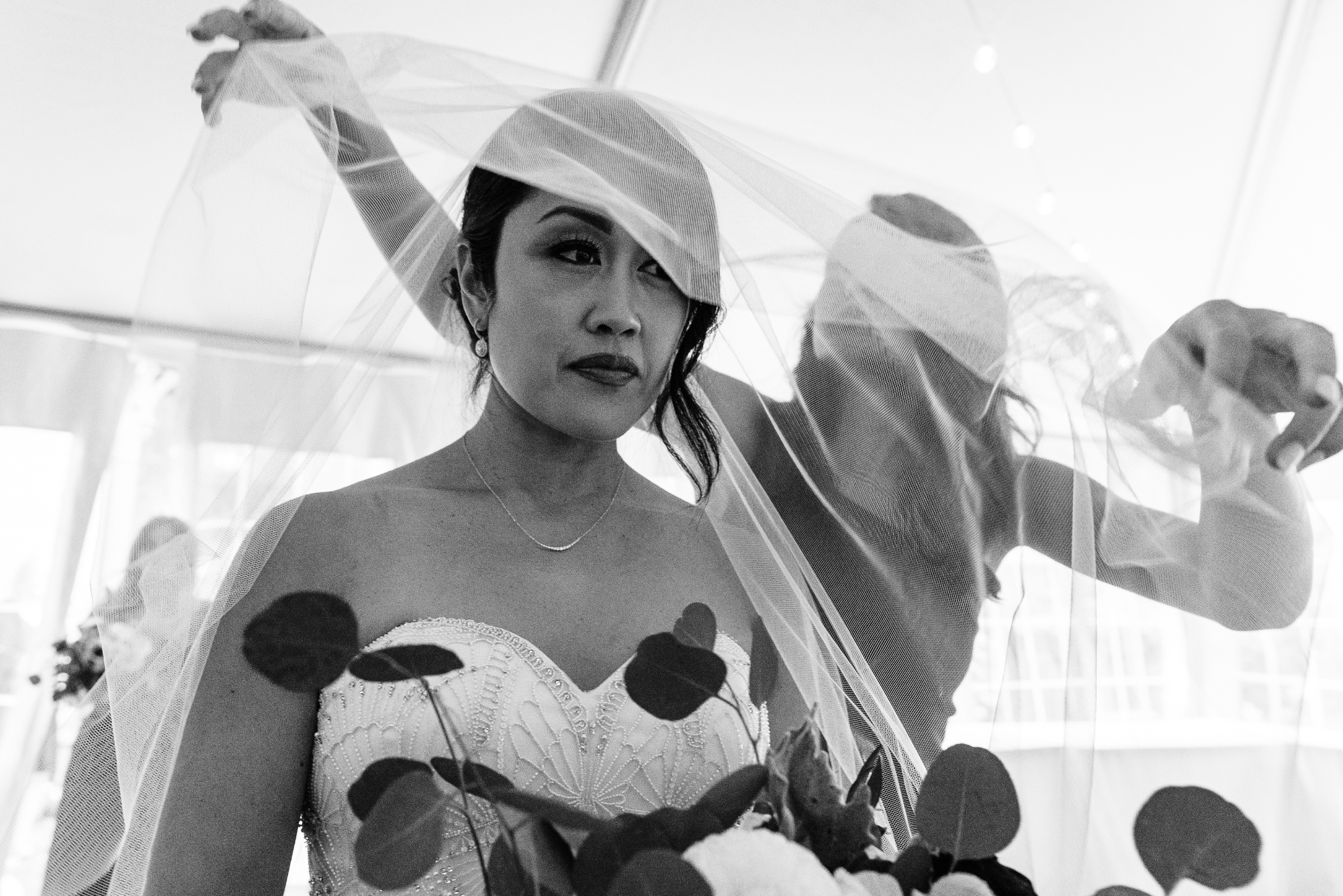 Black and white photo of a bride in a strapless wedding gown, wearing a veil, holding a bouquet, with a woman assisting her in the background.