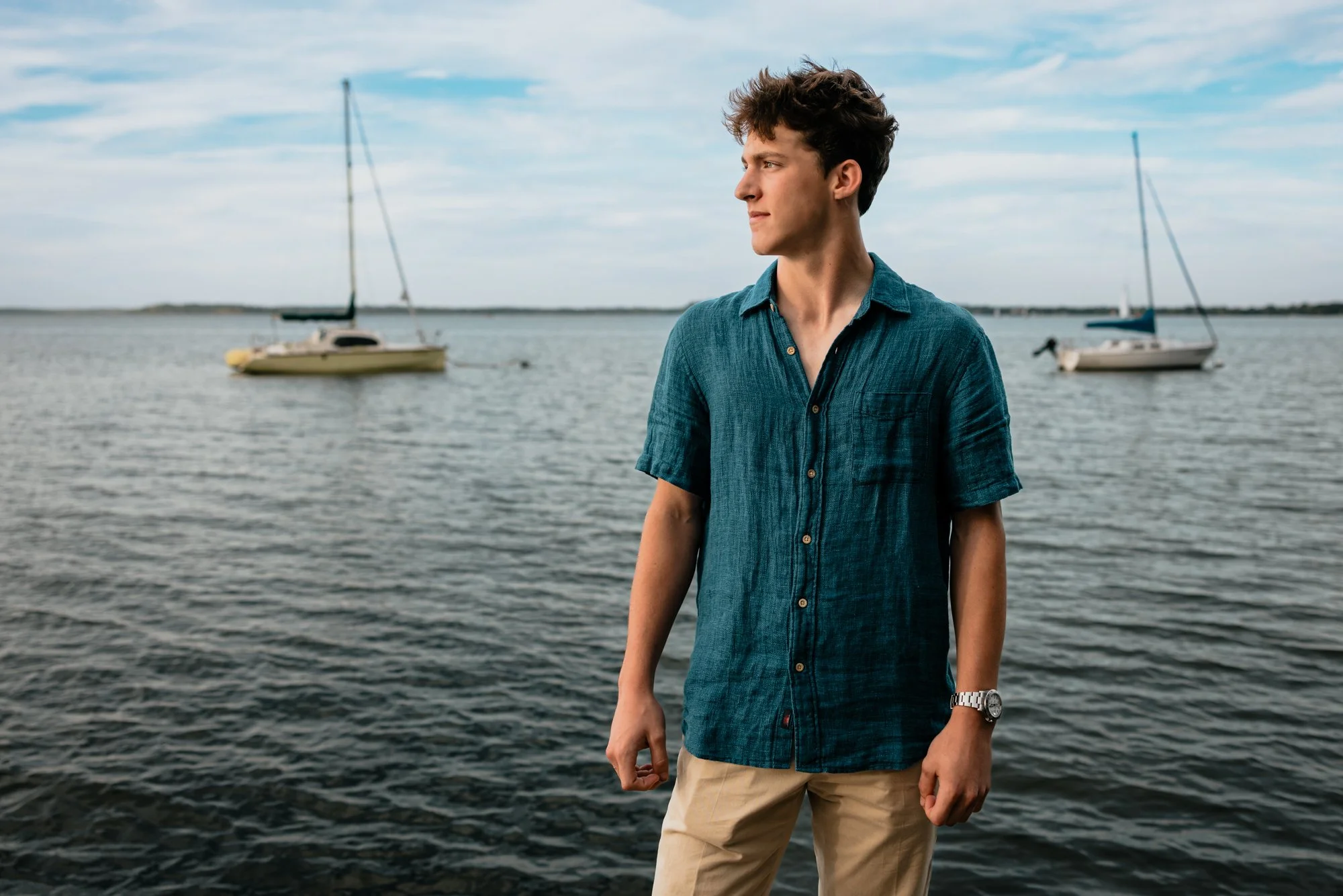 A young man stands by the water with two sailboats in the background, looking to the right.