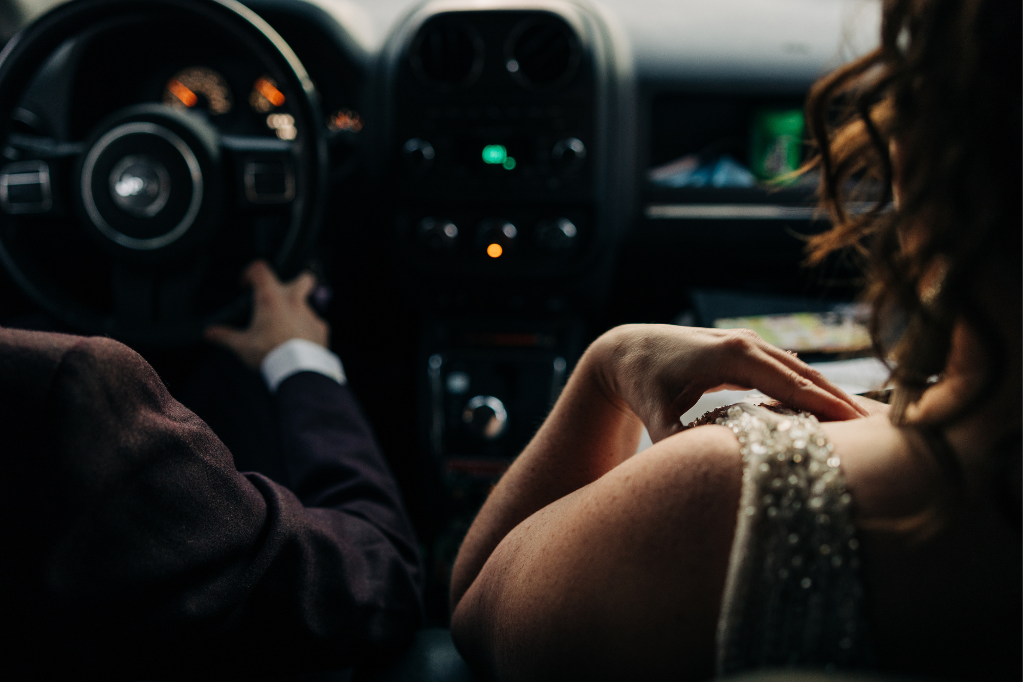 A couple sitting inside a car, with the focus on the woman’s hand resting on her knee, wearing a dress with pearl details, and the man’s hand on the steering wheel. The dashboard and steering wheel are visible in the background.