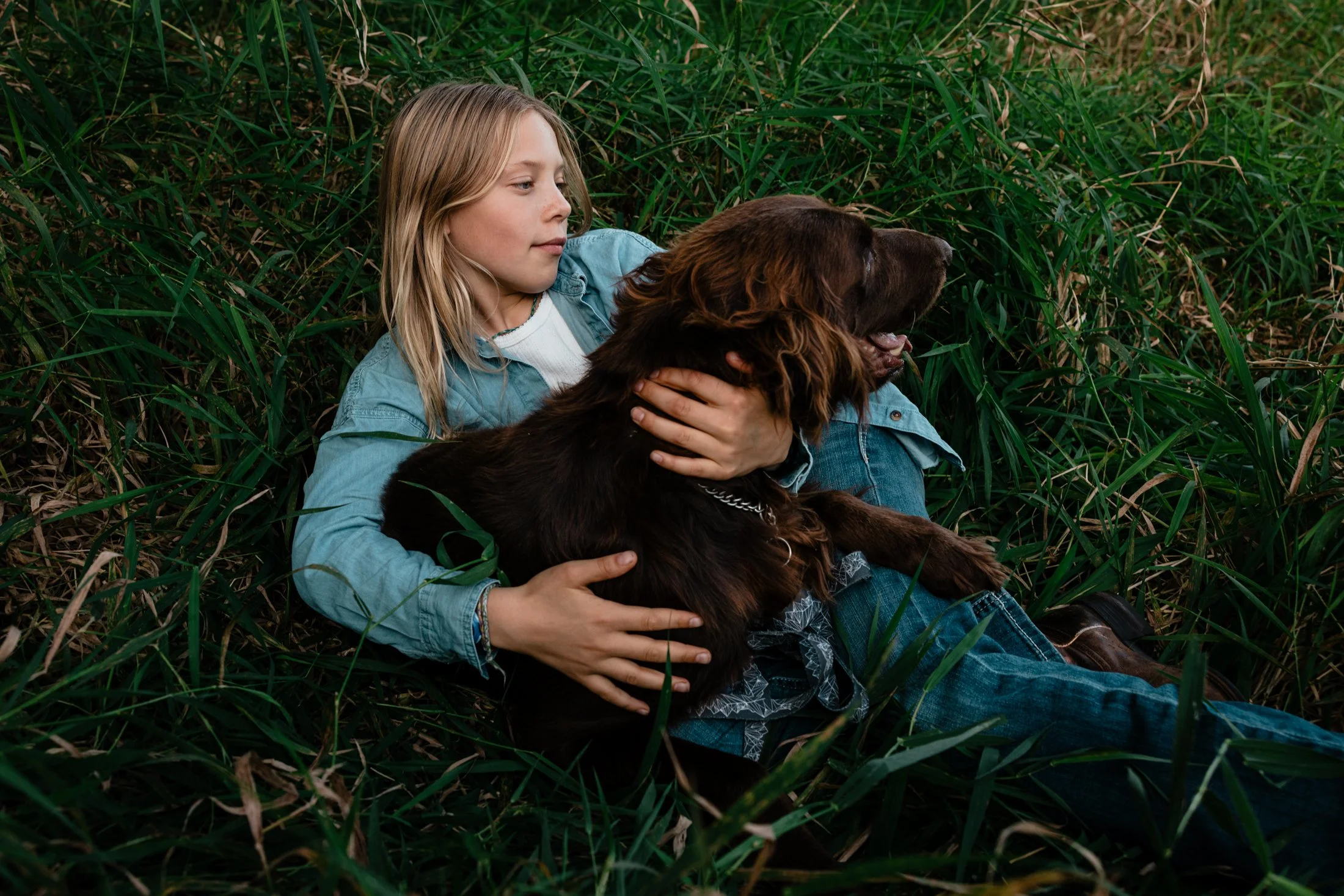 A young girl lying on her side in tall grass, cuddling a brown dog.