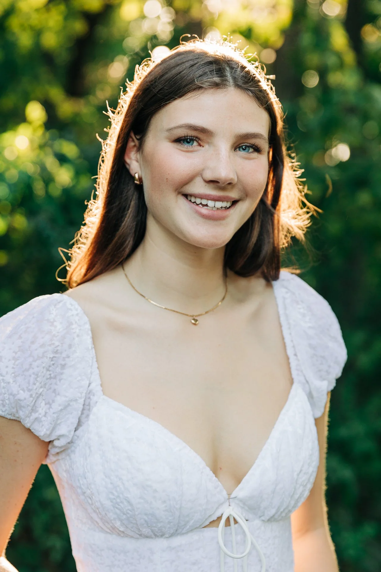 A young woman with long brown hair and blue eyes smiling outdoors in a sunny, green park, wearing a white textured dress with puff sleeves and a gold necklace.