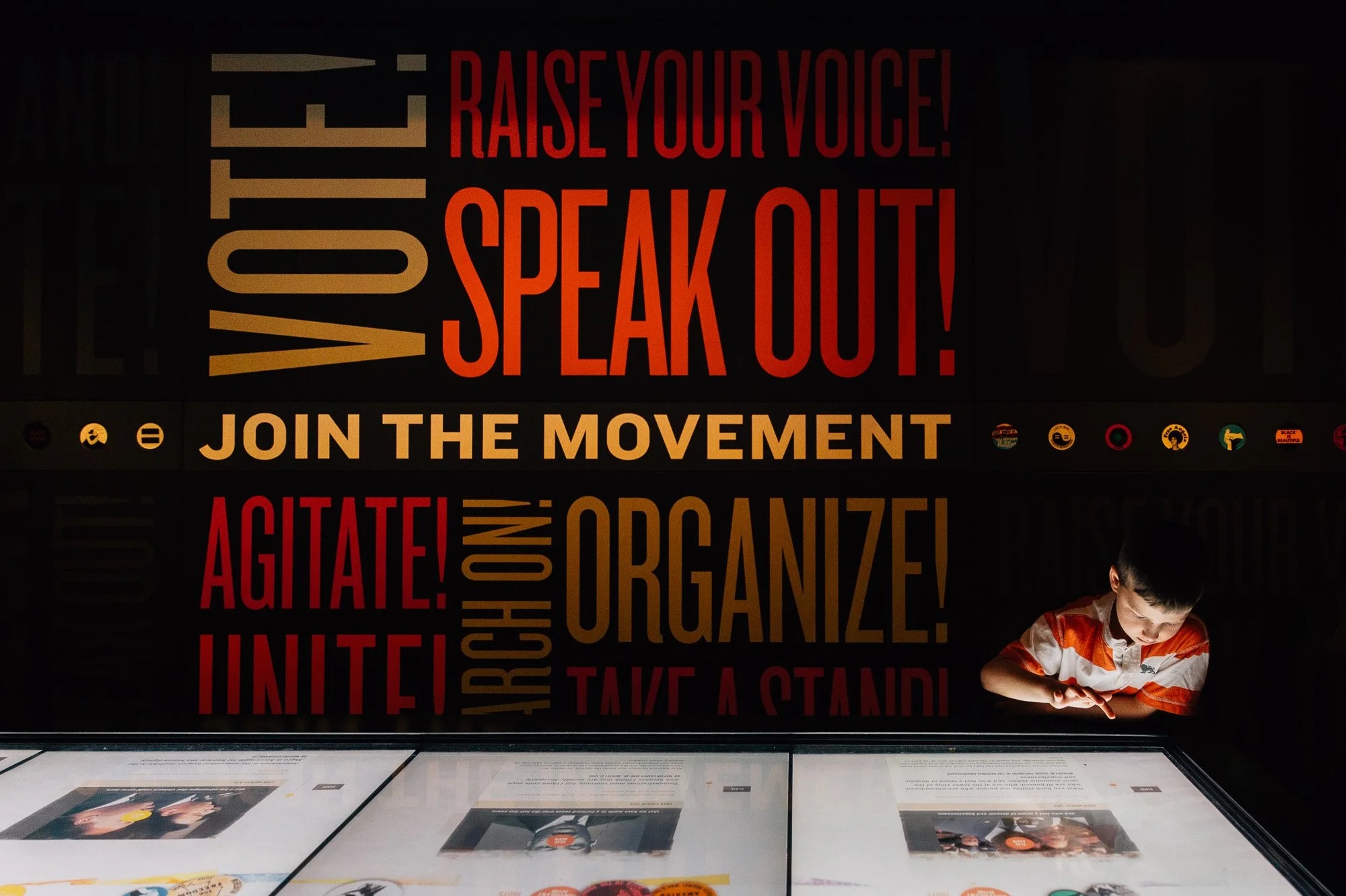 A young boy in an orange and white striped shirt reads a display with bold red and tan text on a black background, encouraging activism and community involvement.