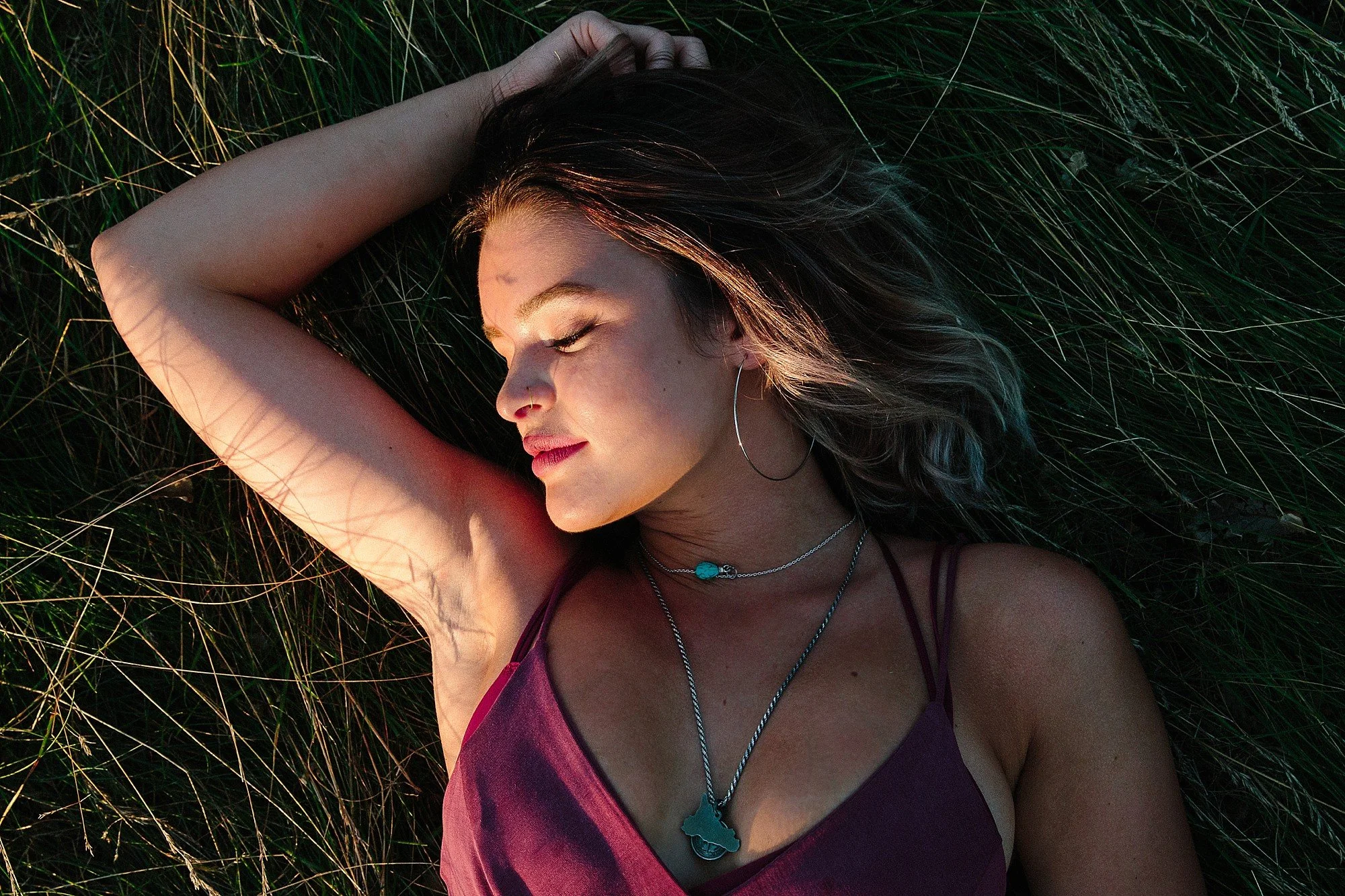 Woman with long dark hair lying on grass with eyes closed, wearing a purple tank top and silver jewelry, including necklaces and hoop earrings.